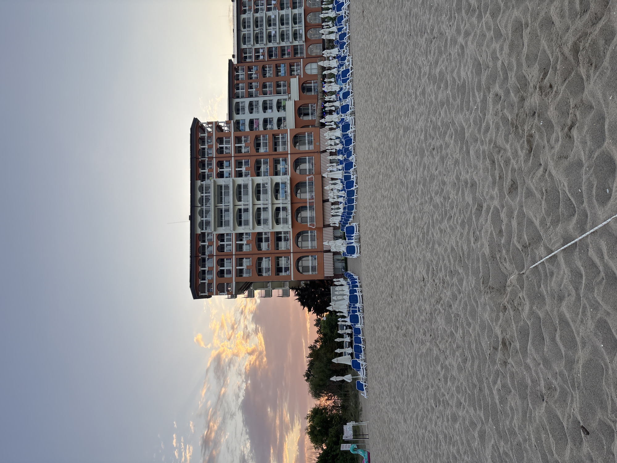 The image depicts a wide view of a sandy beach in Obzor, Bulgaria, at dusk. In the background, a large, multi-story hotel with orange and white facades dominates the scene. The hotel has numerous balconies and arched windows. Rows of blue lounge chairs and white umbrellas are neatly arranged on the sand in front of the hotel, suggesting a resort area. To the left of the hotel, a line of green trees and vegetation is visible. The sky above is a gradient of soft blues and pinks, with wispy clouds tinged with orange from the setting sun. The foreground is filled with textured sand, showing ripples from the wind. A white string or cord is laid across the sand in the lower right portion of the image. No people are visible in the frame.