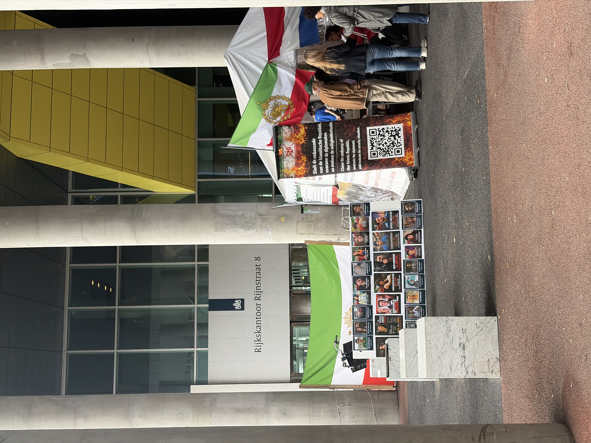 This media file captures an outdoor protest or demonstration taking place in an urban setting, specifically near the "Rijkskantoor Rijnstraat 8" building in The Hague, Netherlands.
The main subjects are several people, partially visible and mostly standing behind protest materials. They appear to be adults, dressed in casual clothing.
Prominently displayed are two flags, both featuring green, white, and red horizontal stripes. One flag, possibly an older Iranian flag, has a golden emblem in its center. Below these flags, a large black banner with orange flames at the top and bottom features text in both Dutch and English, alongside a QR code. The English text reads: "Close the embassy of the Islamic Republic, evacuate the diplomats and evaluate the security situation." The Dutch text conveys the same message: "Sluit de ambassade van de Islamitische Republiek, evacueer de diplomaten en evalueer de veiligheidssituatie." Another smaller, partially visible sign includes "Act Now."
In front of the banners, a grid of numerous portrait-style photographs is displayed, possibly commemorating individuals or calling for justice. Some visible names include "AMIRHOSSEIN HATAMI," "ERFAN SHAKURZADE," "SHAIN VAHEDPARAST," and "AMIRALI MIRJAFARI." Several white marble-like blocks are placed on the ground next to this display.
The setting is an open area with a mix of dark grey and reddish-brown paved ground. In the background stands a modern building with large windows and a distinctive yellow architectural paneling. The building's light-colored facade clearly shows "Rijkskantoor Rijnstraat 8," indicating a government office. Large concrete columns frame the scene. The overall lighting suggests it is daytime, under overcast or diffuse sky conditions. The demonstration conveys strong political sentiment regarding the Islamic Republic, advocating for diplomatic action.