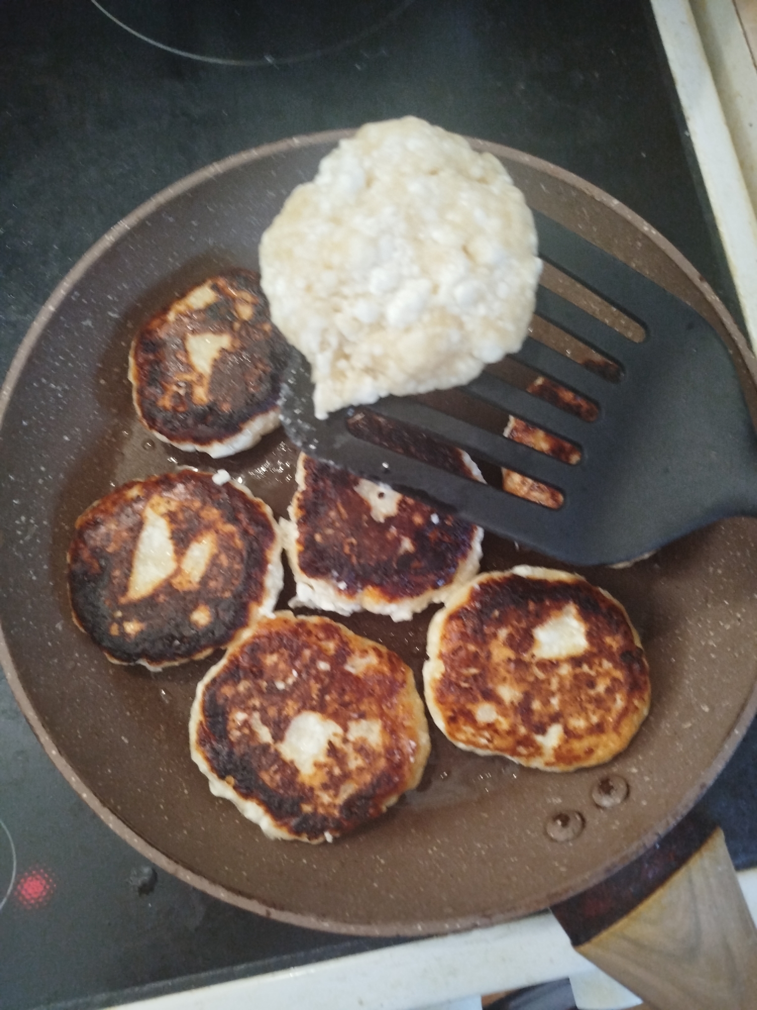 A black frying pan is on a dark stovetop. The pan contains six golden-brown fritters. A black spatula is lifting one of the fritters, revealing a lighter interior. A portion of the fritter on the spatula is slightly burnt. The stovetop appears to be electric, with a red heating element visible in the lower left corner. The scene is likely indoors, in a kitchen.