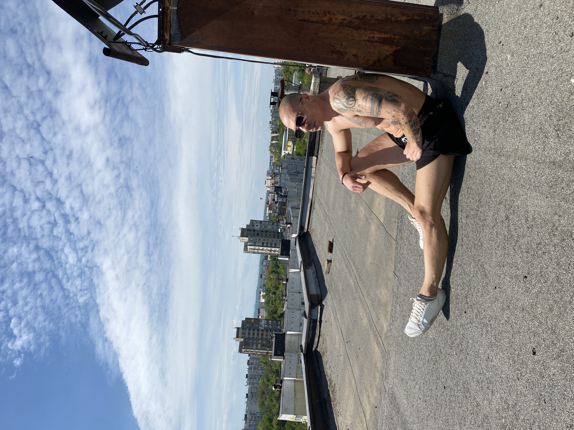A man with a shaved head and tattoos on his arms is kneeling on a flat rooftop in an urban environment. He is wearing sunglasses, black shorts, and white sneakers. Buildings are visible in the background. The sky is partly cloudy. It is not possible to confirm the location is in the city of Pleven, Bulgaria based on the imagery alone.