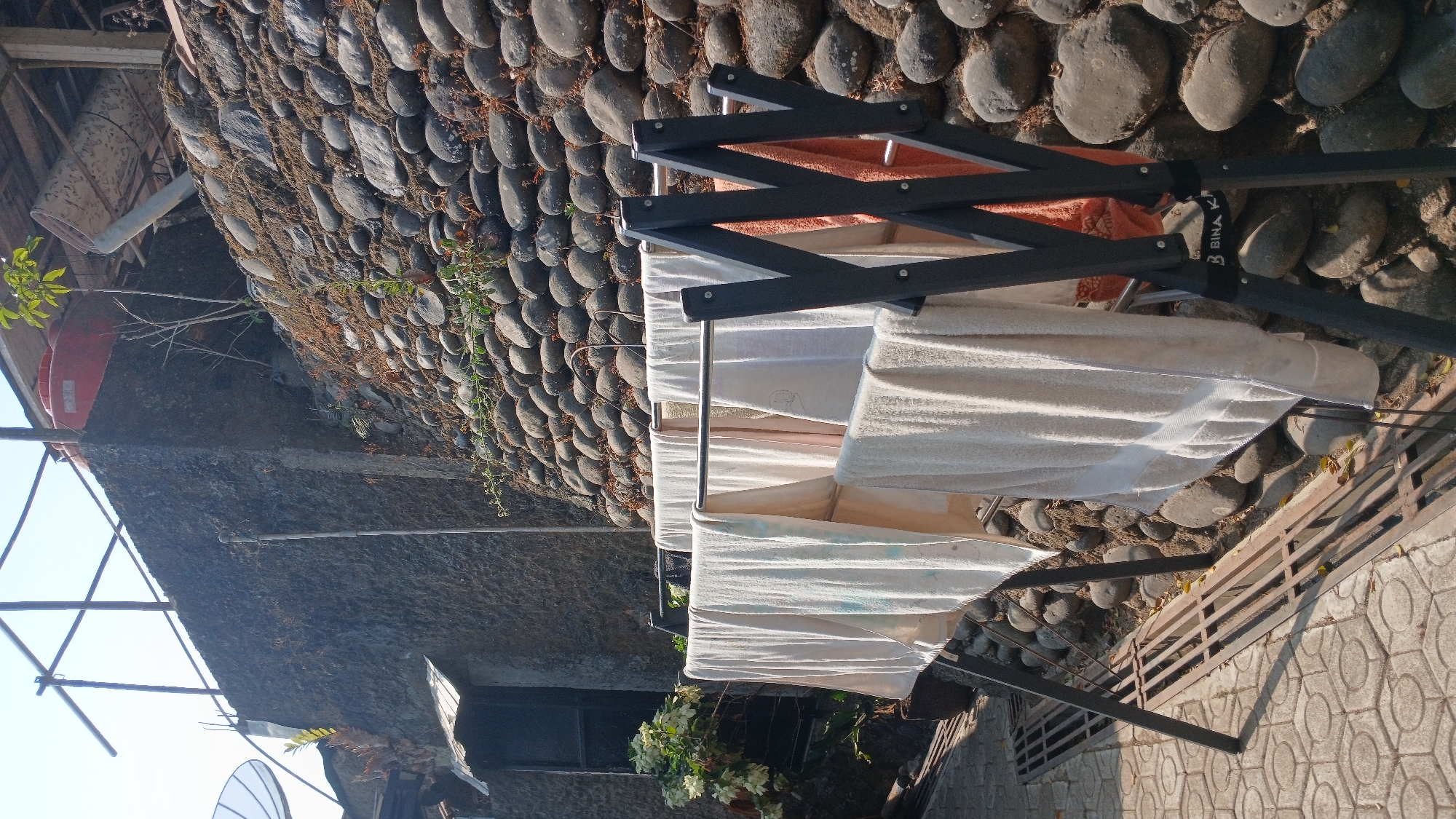 A clothes drying rack with several white towels and one orange towel hangs in the foreground, positioned on a paved area with a metal grate next to it. Behind the drying rack is a textured concrete wall, which transitions into a sloping wall constructed of rounded river stones. A small tree with sparse green leaves grows from the stone wall. In the upper left corner, a portion of a white satellite dish is visible. Further up, a red water tank sits atop the concrete structure. The scene appears to be an outdoor residential area, possibly in a hilly or terraced landscape, bathed in bright sunlight. The city of Sampang, Indonesia, is known for its coastal areas and hilly terrain, making such a scene plausible within its context.