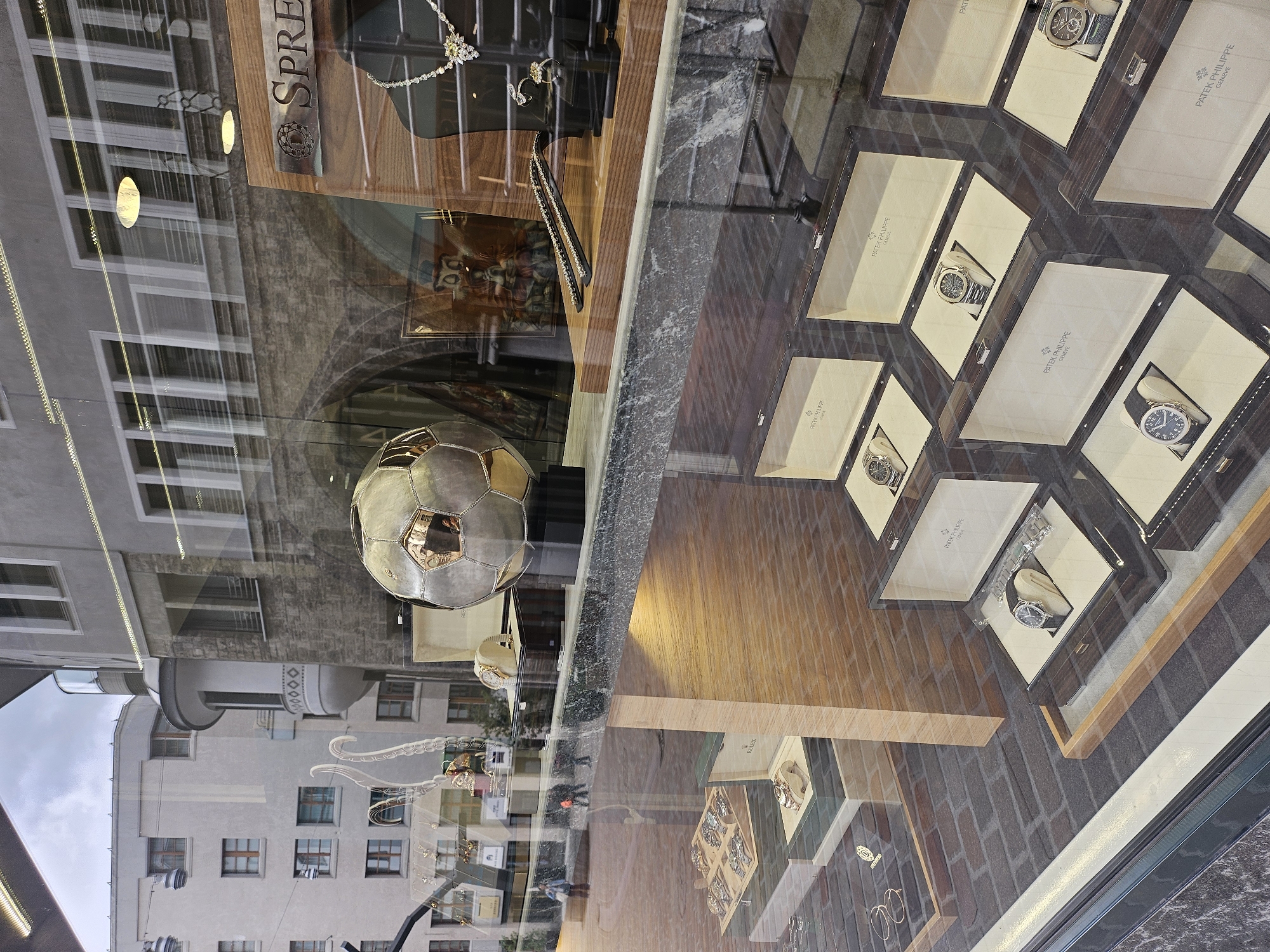This image shows a street-level view of a luxury watch and jewelry store's display window, with reflections of the street and buildings visible. The primary subjects are the watches displayed in individual boxes, most of which are branded "PATEK PHILIPPE GENEVE" and one is branded "ROLEX". There are also necklaces and rings visible in the display. A large, metallic soccer ball is prominently featured, reflecting the surrounding environment. The scene is likely set in a city, given the architecture of the reflected buildings, and the presence of a store suggests a commercial area. The lighting suggests it could be daytime. There is text visible on the watch boxes and on a sign above the display, reading "ROLEX" and "SPRE". The overall impression is one of affluence and high-end retail.