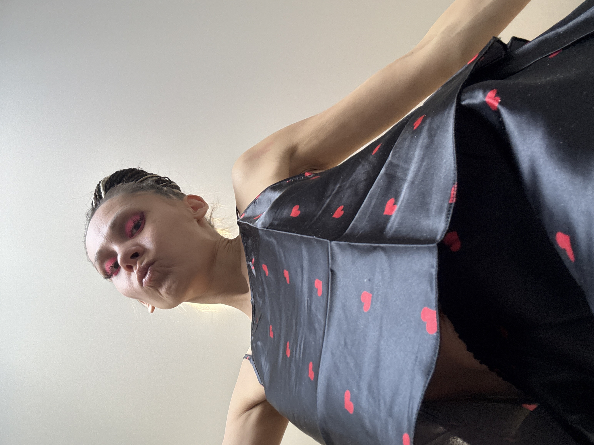 A woman with braided hair and pink eyeshadow is looking down at the viewer with pursed lips. She is wearing a black dress with red heart patterns. The background is a plain white wall or ceiling. The photograph is taken from a low angle, looking upwards towards the woman. The location is not discernible from the image.