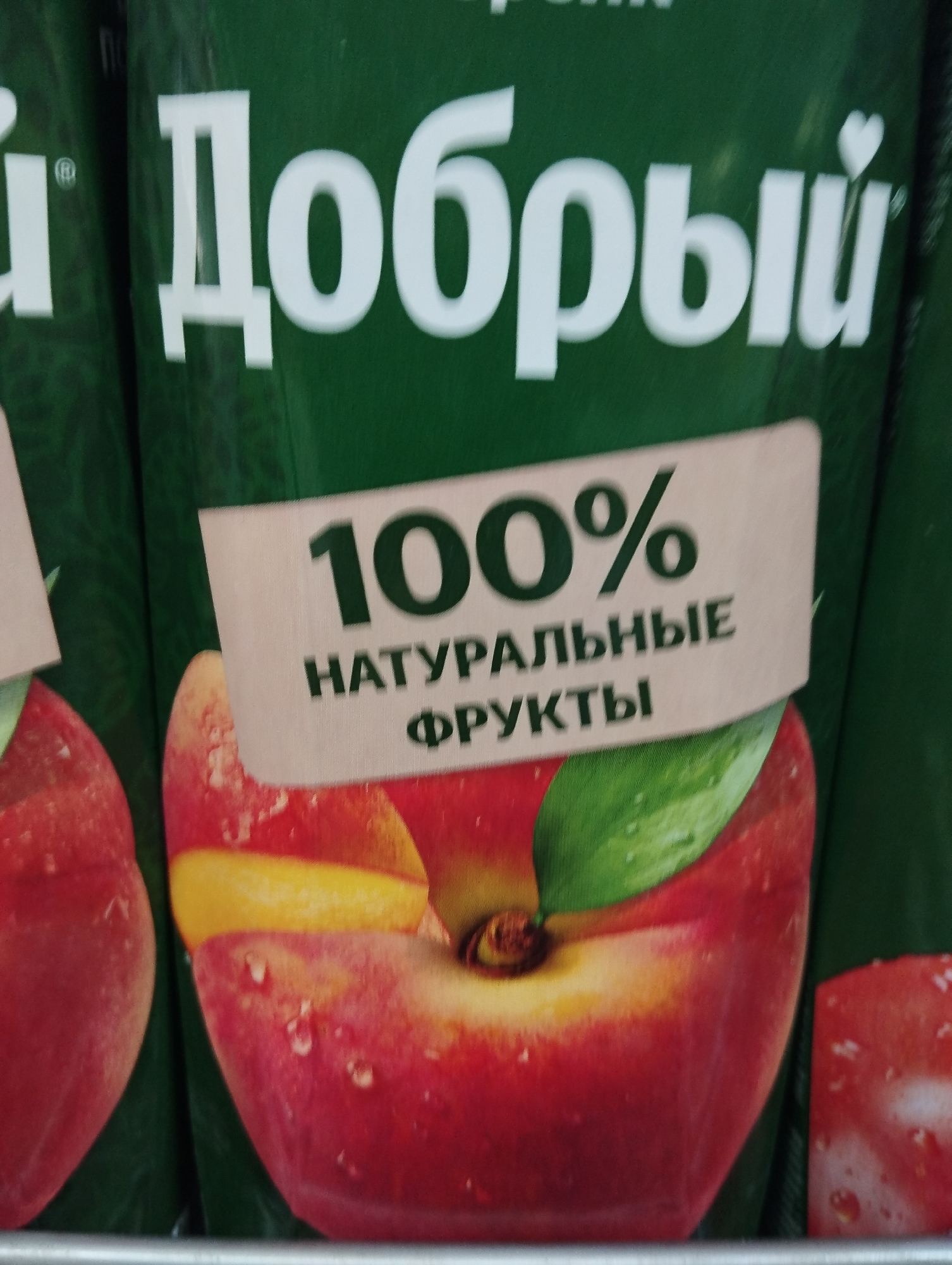 Close-up shot of a green juice carton with white Cyrillic text "Добрый" (Dobryi) prominently displayed on top. A cream-colored sticker partially covers the carton with black text stating "100%" and "НАТУРАЛЬНЫЕ ФРУКТЫ" (NATURAL FRUITS) in smaller letters beneath. The sticker also depicts a red and yellow apple with water droplets and a green leaf. Another similar juice carton is partially visible on the left side of the frame. This photo was taken in Krasnogorskoye, Russia.