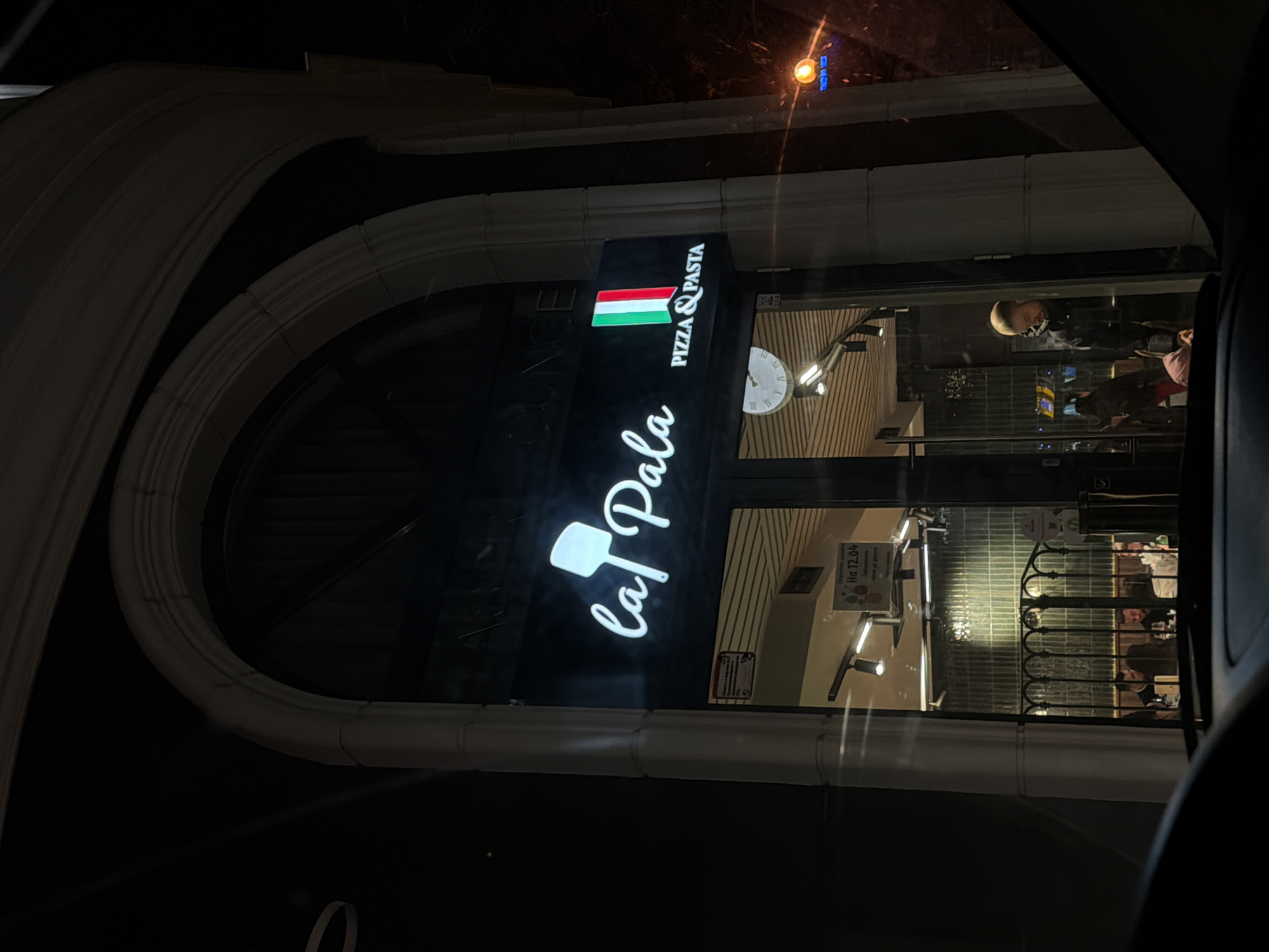 The image shows the exterior of "La Pala," a pizza and pasta restaurant in Varna, Bulgaria, at night. The restaurant's sign features a logo with a pizza paddle and the colors of the Italian flag. Through the glass facade, tables and chairs, metal rails, and several individuals are visible inside. A clock hangs on the wall. Light fixtures illuminate the interior. The architectural style of the building includes a prominent arched doorway.