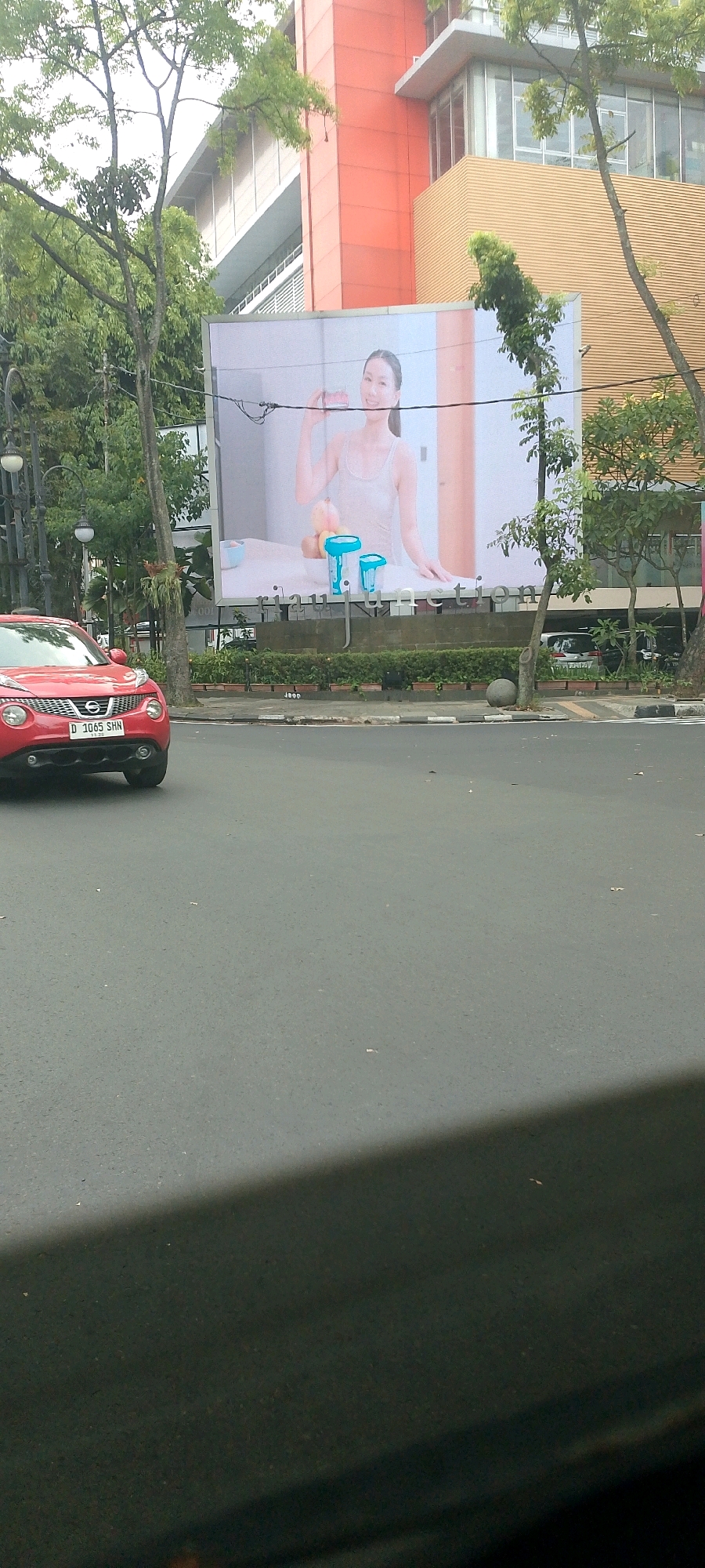 In Bandung City, Indonesia, a red Nissan Juke car with license plate D 1065 SHN is captured in motion on an asphalt road. The car is positioned on the left side of the frame, moving away from the viewer. Behind the car, a large billboard is prominently displayed, featuring a woman in a light-colored tank top holding a pink object. The billboard is situated in front of a building with orange and yellow architectural elements, some of which are adorned with glass windows. Lush green trees and foliage surround the billboard and the building, indicating a well-vegetated urban environment. The sky is overcast. A shadow, possibly from a vehicle or structure, stretches across the lower portion of the image.
