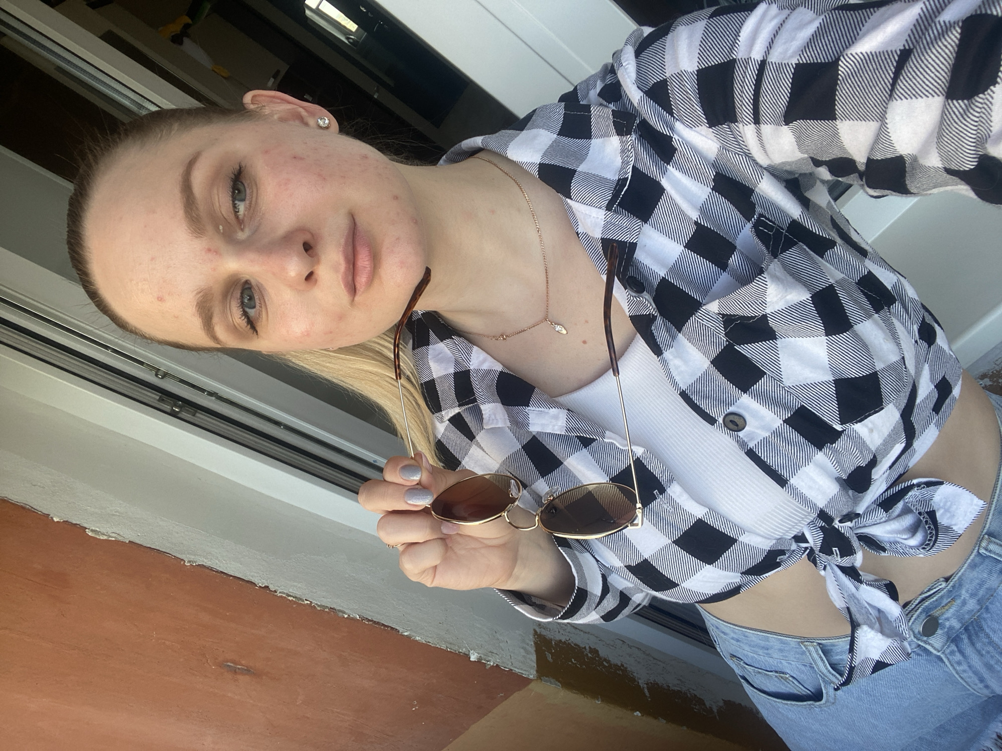 A young woman with blonde hair tied back in a ponytail stands near a window. She is wearing a white tank top, a black and white plaid shirt tied at the waist, and blue denim shorts. She is holding a pair of sunglasses. The background shows a building with a textured orange wall and a white window frame. The woman has acne on her face and is wearing a silver stud earring and a delicate gold necklace.