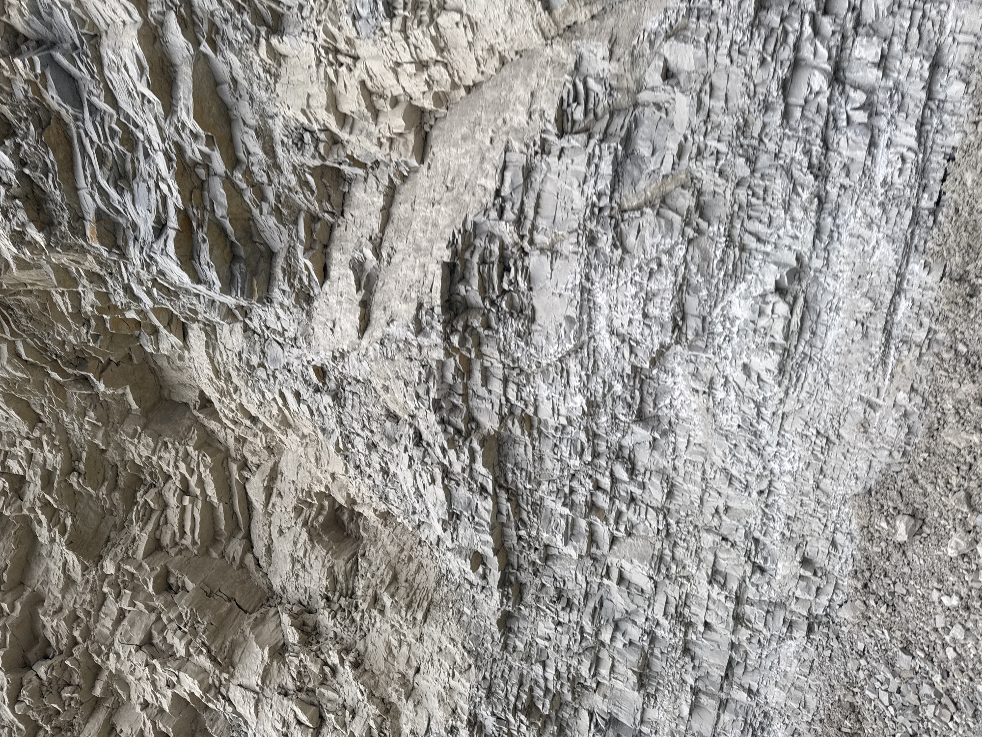 This is a close-up, oblique, eye-level shot of a layered rock face, likely a cliff or excavation, exhibiting distinct horizontal striations. The rocks are predominantly gray and tan, with varying degrees of fragmentation and texture, suggesting sedimentary rock. Some areas show signs of erosion, with smaller pebbles and debris accumulated at the base. The lighting is overcast, resulting in a diffused, shadowless illumination across the surface. No people or other discernable subjects are present. The image depicts a natural geological formation.