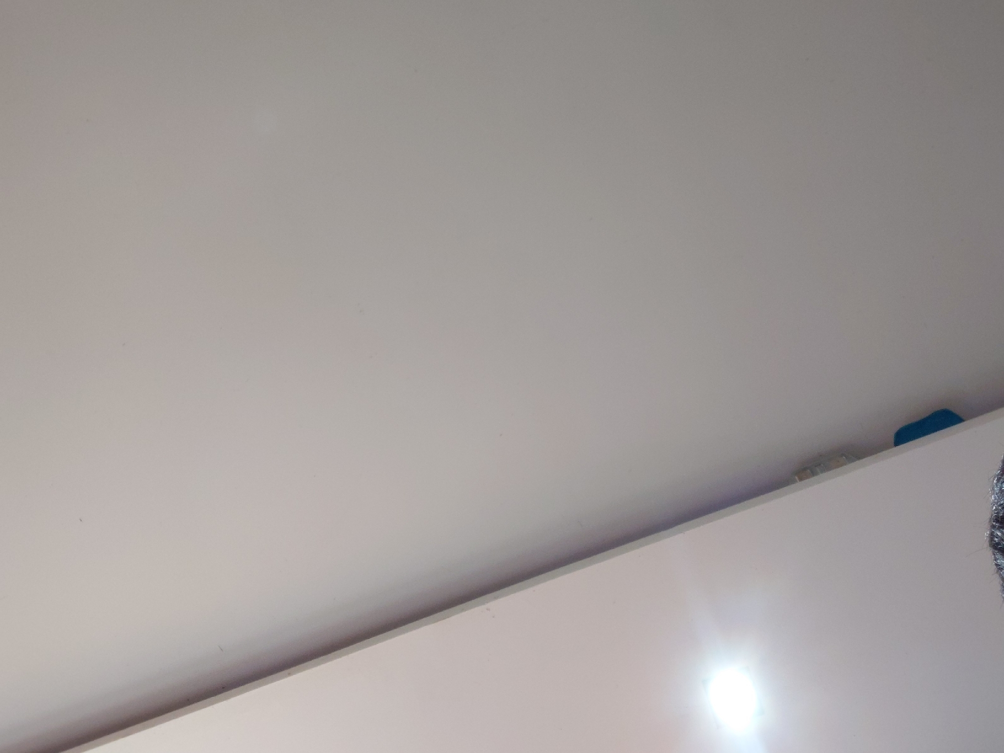 The image displays an upward-angled indoor view, predominantly featuring a light grey or off-white ceiling. The ceiling is a flat, uniform surface, occupying the upper and central portions of the frame. A distinct, slightly shadowed horizontal line runs diagonally across the lower part of the image, marking the boundary where the ceiling meets a perpendicular white surface, likely the top edge of a wall or a tall piece of furniture such as a cabinet or shelf.
On this white surface, in the lower right quadrant, there is a very bright, circular reflection of a light source, indicating a well-lit environment. On the top edge of the white furniture, in the upper right corner, a small blue object is visible, along with another indistinct object behind it. On the extreme right side of the frame, a sliver of a dark, textured object with a slight metallic sheen is barely visible, suggesting its presence just outside the frame.
The setting is an interior space in Santa Ana, Venezuela. No specific activity or event is discernible, and no text is visible. The bright lighting suggests it could be daytime or an area with strong artificial illumination.