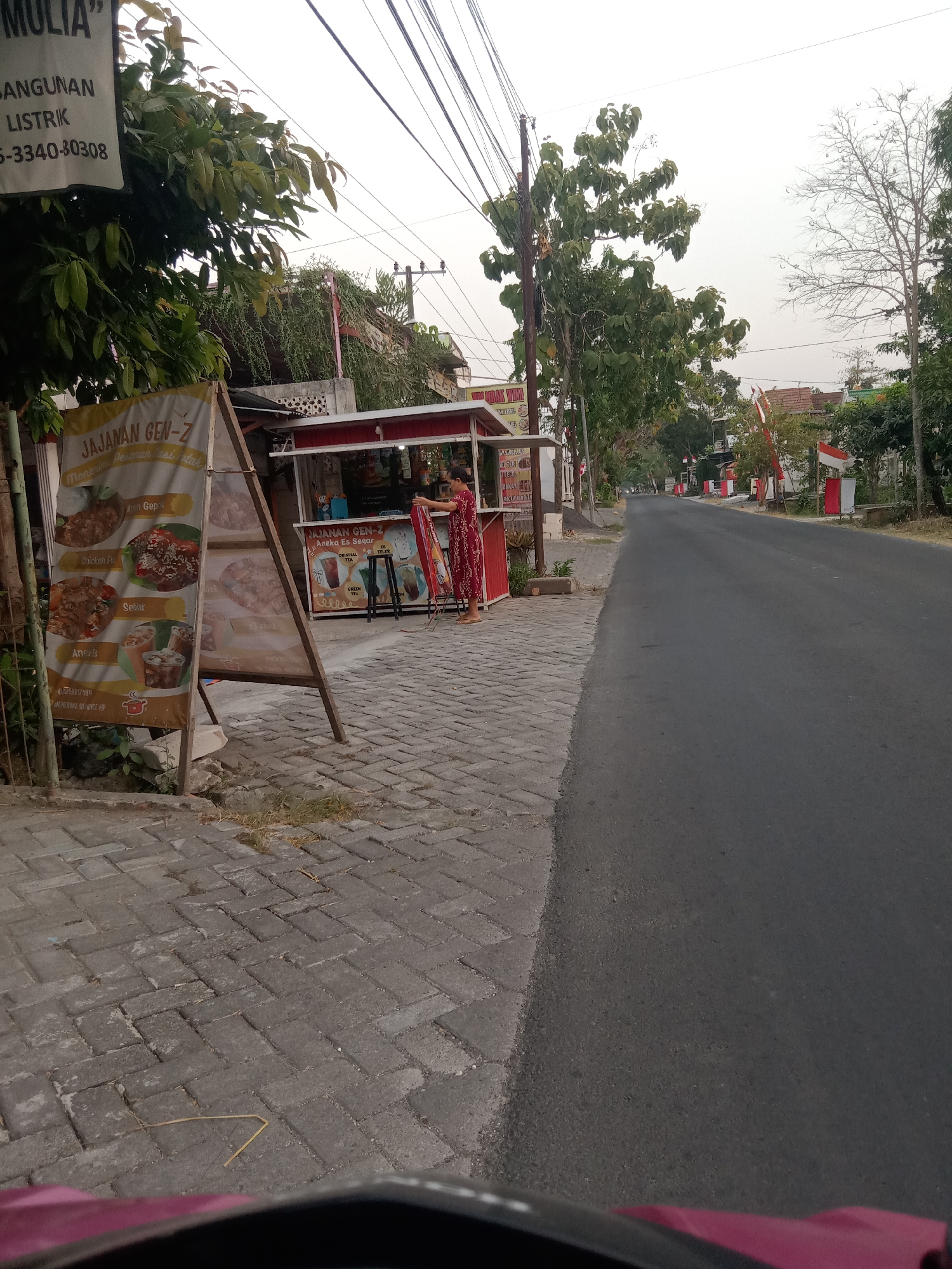 The image shows a street scene in Sugihwaras, Indonesia. On the left side of the street, there is a food stall with an A-frame sign displaying pictures of food items. Next to the stall is a woman in a red dress who appears to be arranging a banner. A paved sidewalk with a brick pattern runs along the street. The street itself is paved and appears to be a main road, with trees and utility poles lining the sides. In the background, there are buildings and more trees. The sky is overcast, suggesting it might be a cloudy day. The overall impression is that of a typical street in a residential or commercial area of an Indonesian town.