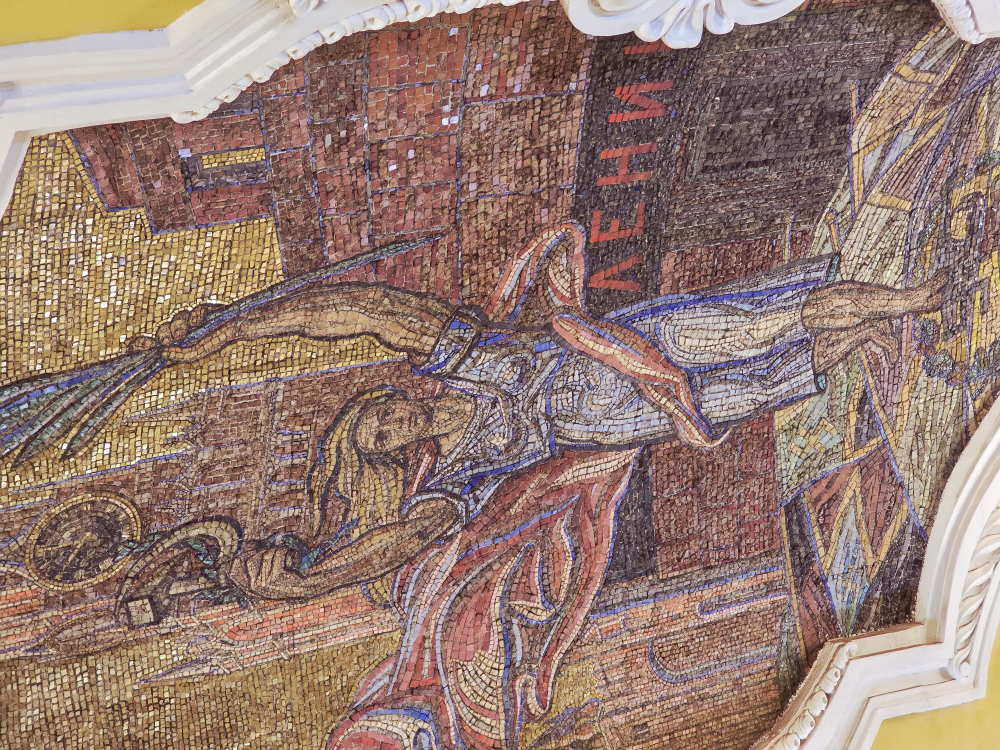 The image shows a mosaic mural, likely located in Moscow, Russia. The mural depicts a woman with long, flowing hair, possibly a mythological or allegorical figure, seemingly being transported or carried. She is draped in clothing or fabric colored in shades of red, blue, and white. She appears to be holding a staff or similar object in one hand and is reaching towards the left of the frame. The background is a mosaic of varying shades of brown, gold, and dark red, resembling a city or landscape scene. The overall style suggests a Soviet-era artistic aesthetic, possibly depicting a scene of labor, progress, or national identity. The mosaic is bordered by a white, ornate architectural molding, indicating that it is located on a ceiling or wall within a building. The text "ЛЕНИН" (LENIN) is visible. The subject is located at the Kievskaya metro station in Moscow.
