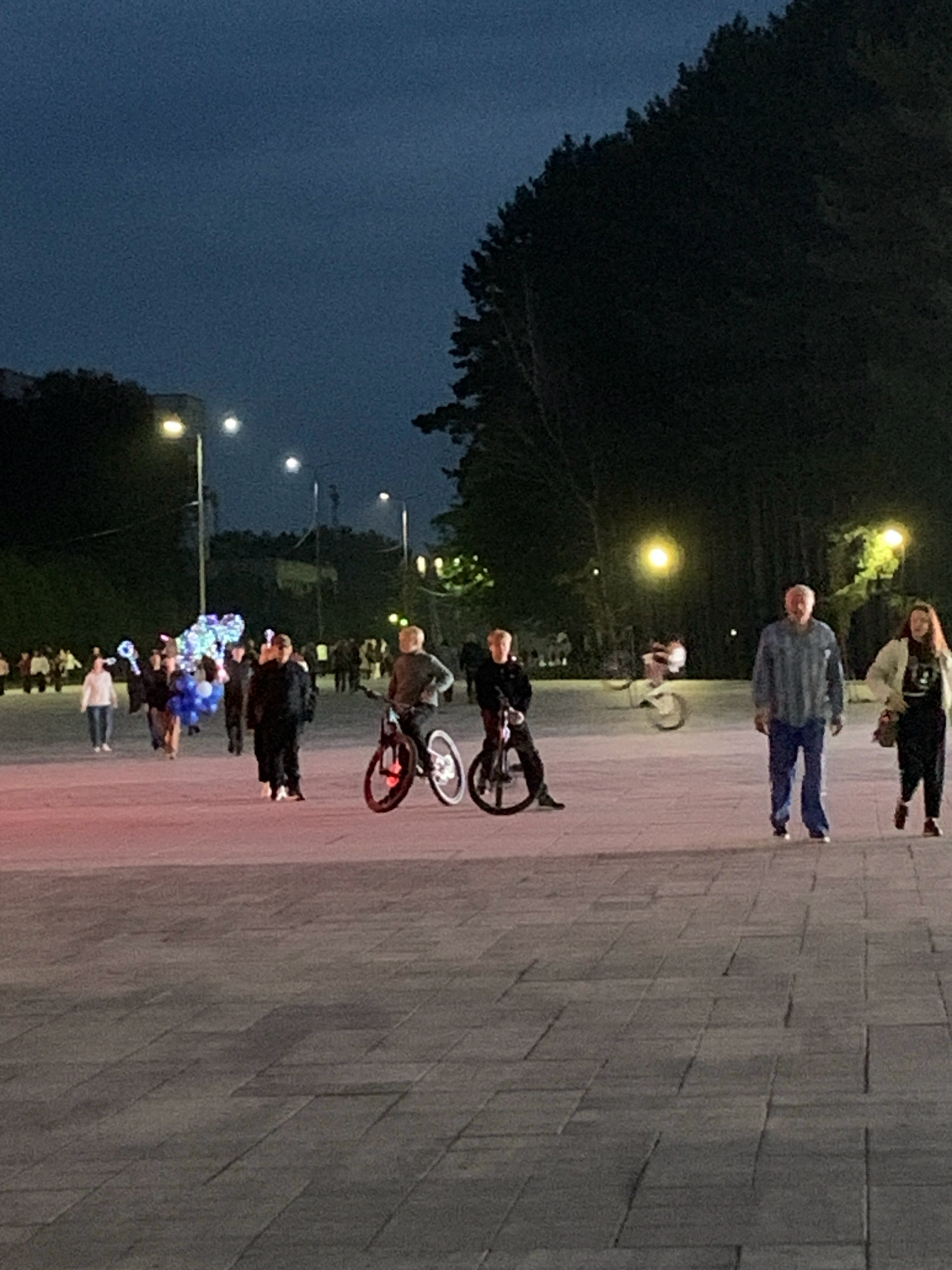 The image depicts an outdoor scene at dusk, likely in a public park or pedestrian area in Prokopyevsk, Russia. In the foreground, a wide, paved surface with distinct stone tiling is visible. Several individuals are present, engaged in various activities.
On the right side of the frame, an adult male wearing a blue plaid shirt and blue pants is walking towards the viewer, accompanied by an adult female in a white jacket over a dark outfit. To the left of them, a young person on a bicycle is also in motion.
Further to the left, two individuals are positioned on bicycles. One is a younger person in dark clothing on a bicycle with illuminated red rims. Another person, seen from the back, is on a bicycle with a red and white frame. In the mid-ground, a group of people are walking, some holding what appear to be illuminated balloon decorations, predominantly blue and white.
In the background, streetlights illuminate the area, casting light on pathways and trees. The sky is dark, indicative of evening. The overall atmosphere suggests a public gathering or event taking place in an urban setting.