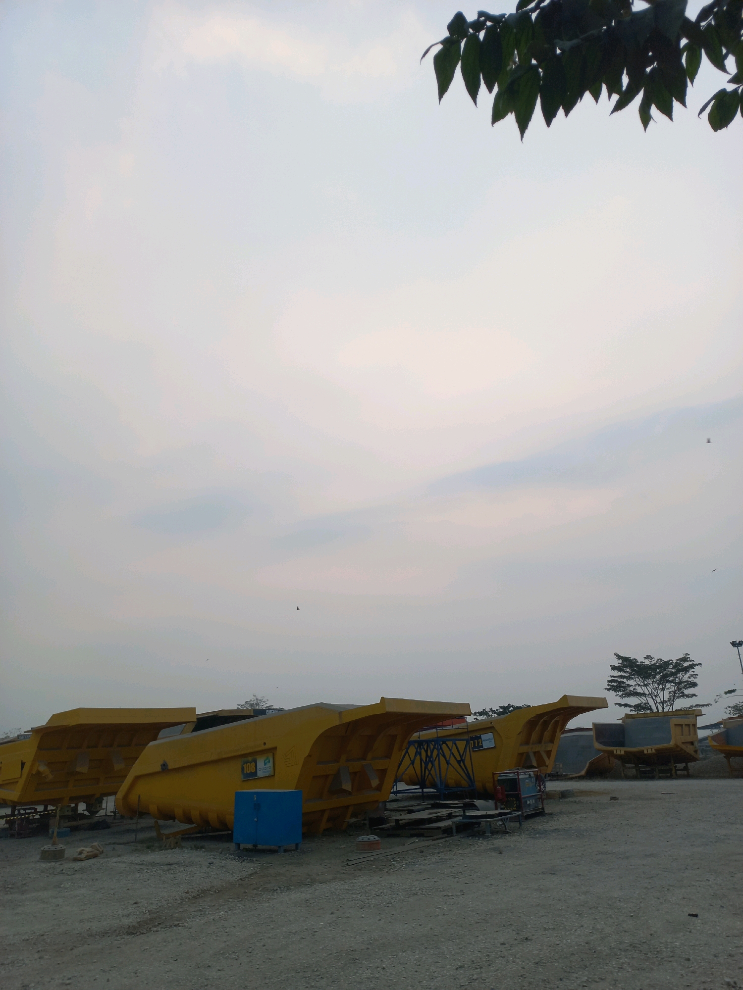 In Paringin, Indonesia, a lot of large yellow dump truck beds are parked on a gravel lot. The sky above is overcast and pale. In the foreground, the truck beds are lined up, some with visible numbers like "108" and "112." There are blue storage containers and metal scaffolding near the truck beds. In the background, there are trees and what appear to be more dump trucks or piles of earth. The overall scene suggests a construction or manufacturing site.