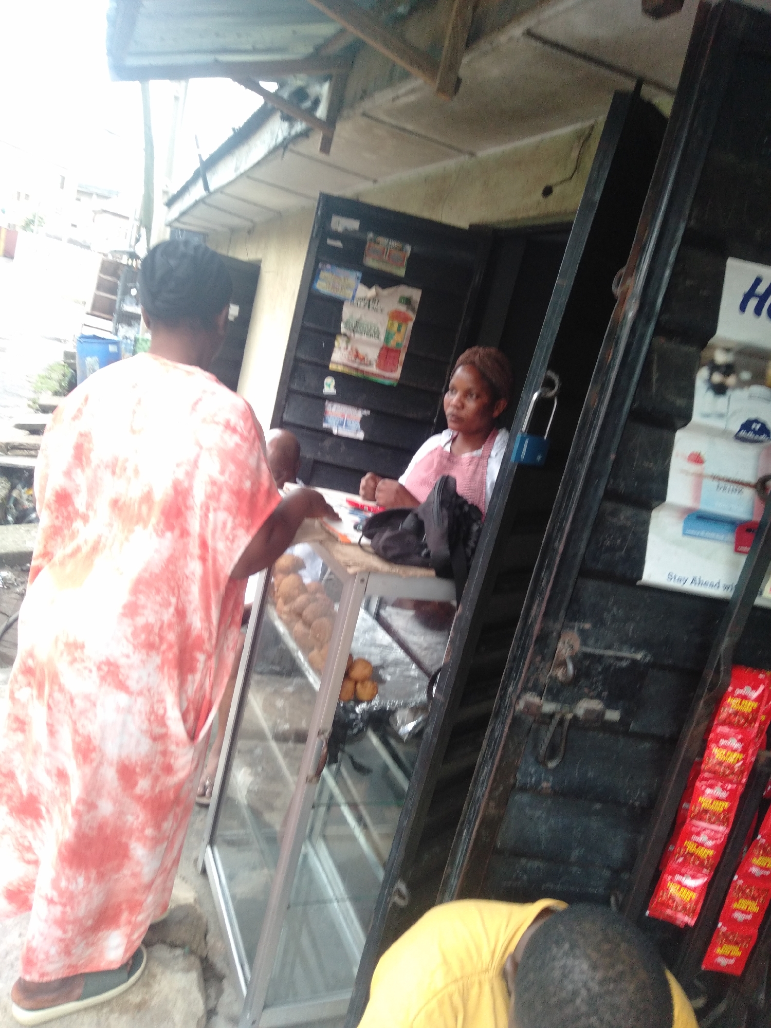 A street scene in an unidentified city. A woman wearing a pink and white tie-dye garment is at a glass-fronted display cabinet in front of a small shop. Inside the shop, a second woman in a pink shirt and headband sits behind the counter. A black backpack is on the counter. The shop's exterior is weathered, with promotional signs and packaged goods affixed to its side. A third person, wearing a yellow shirt, is partially visible in the lower part of the picture.