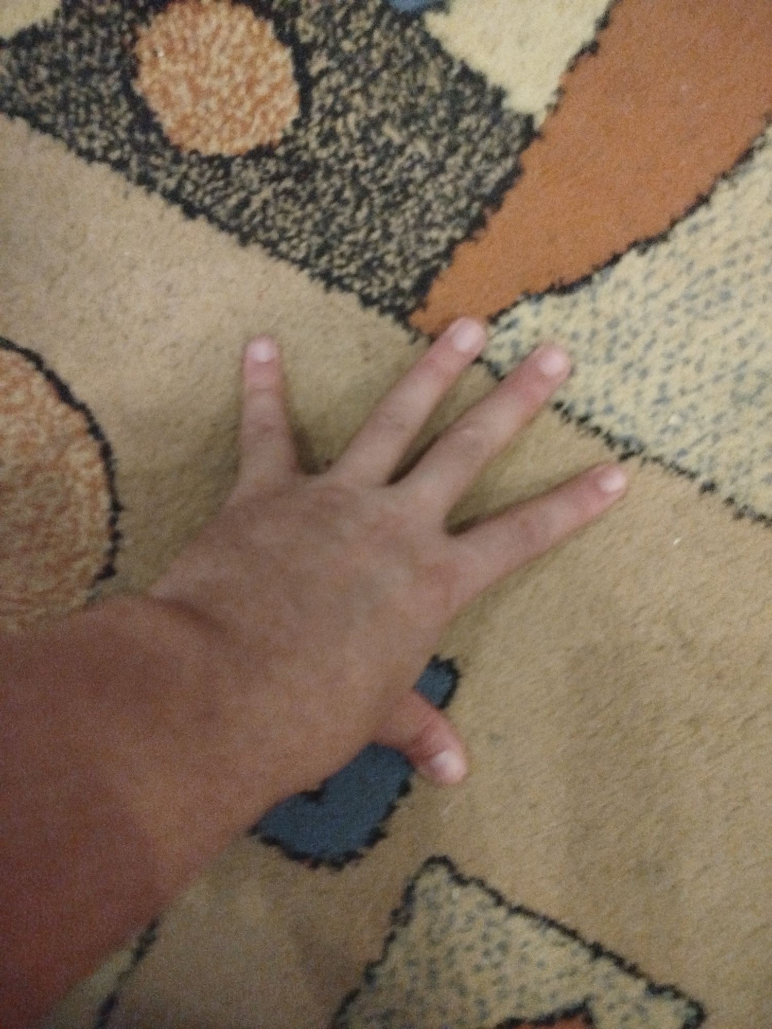 A human hand, with visible fingers and fingernails, is positioned on a carpet. The carpet has a patterned design with various shapes and colors, including tan, brown, orange, black, and blue. The hand is light-skinned. There is no discernible information about the location in Votkinsk, Russia. The image is a close-up shot focusing on the hand and the carpet.