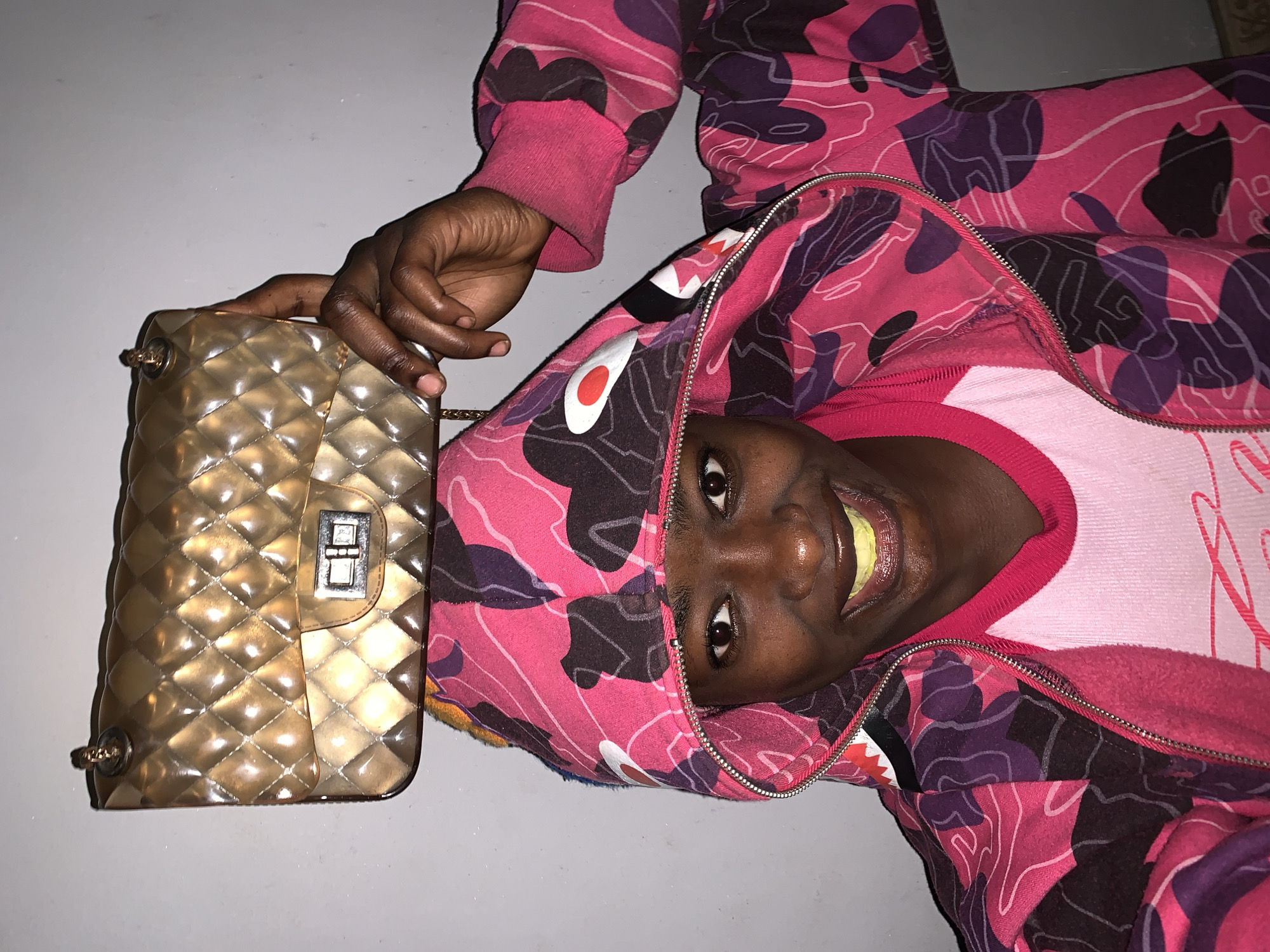 A dark-skinned person wearing a pink and purple camouflage hooded sweatshirt and a light pink crew-neck shirt lies on a light gray surface. The person is holding a tan quilted handbag with a metallic clasp. The location is unknown.