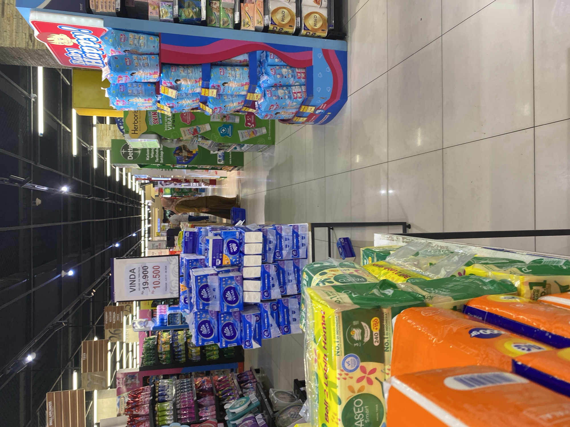 The image shows a retail environment, likely a supermarket or department store, with aisles of merchandise. In the foreground, stacks of packaged facial tissues are visible, with branding like "Paseo" and orange packaging. Further back, on the right, a display of "Baby Happy!" brand diapers is prominently featured. To the left, shelves are stocked with various cleaning and personal care products. A sign labeled "VINDA" with pricing information is also visible. A woman wearing a hijab is seen walking in the background, and other shoppers are present but not clearly identifiable. The store appears to be well-lit, with a tiled floor and a suspended ceiling with lighting fixtures. The presence of Indonesian language on signs and product packaging indicates the location is likely Indonesia. Given the specific product branding and store layout, this scene could be a supermarket in Banda Aceh, Indonesia, such as a Hypermart or similar large-format retail store.