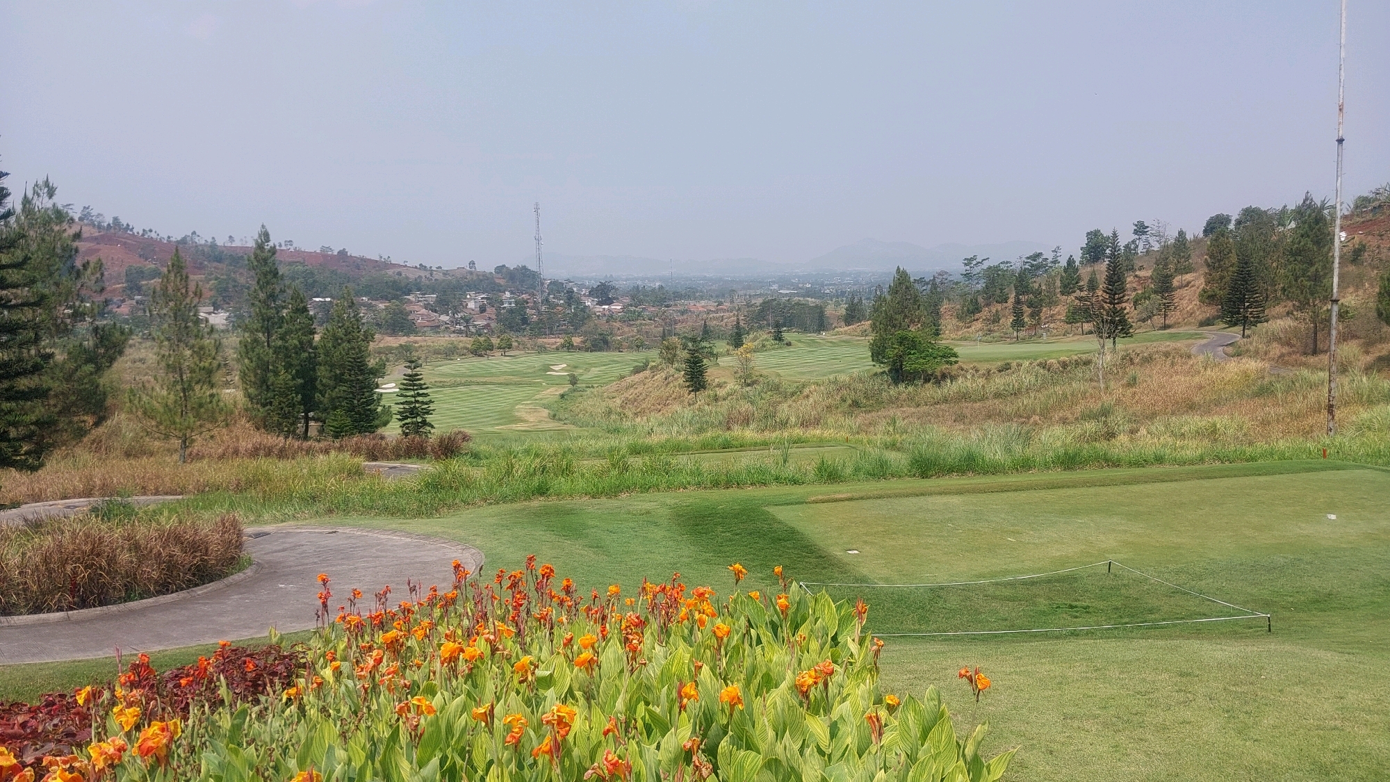 The image captures a wide panoramic view of a golf course nestled in the hilly terrain surrounding Cikande, Indonesia. In the foreground, a vibrant display of orange canna lilies and other green foliage creates a colorful border along a winding paved path. Beyond this, the manicured greens of the golf course stretch out, marked by white boundary lines. The undulating landscape is dotted with various trees, including tall pine trees, and interspersed with patches of dry, golden grass. In the mid-ground, a golf cart path snakes through the course, and sand traps are visible on some holes. In the distance, a hazy cityscape is spread across the valley, with buildings and houses nestled amongst the hills. A prominent telecommunication tower stands tall on a distant hilltop, dominating the skyline. The sky is a pale, hazy blue, suggesting a clear but perhaps warm day. There are no people or animals visible in the image.