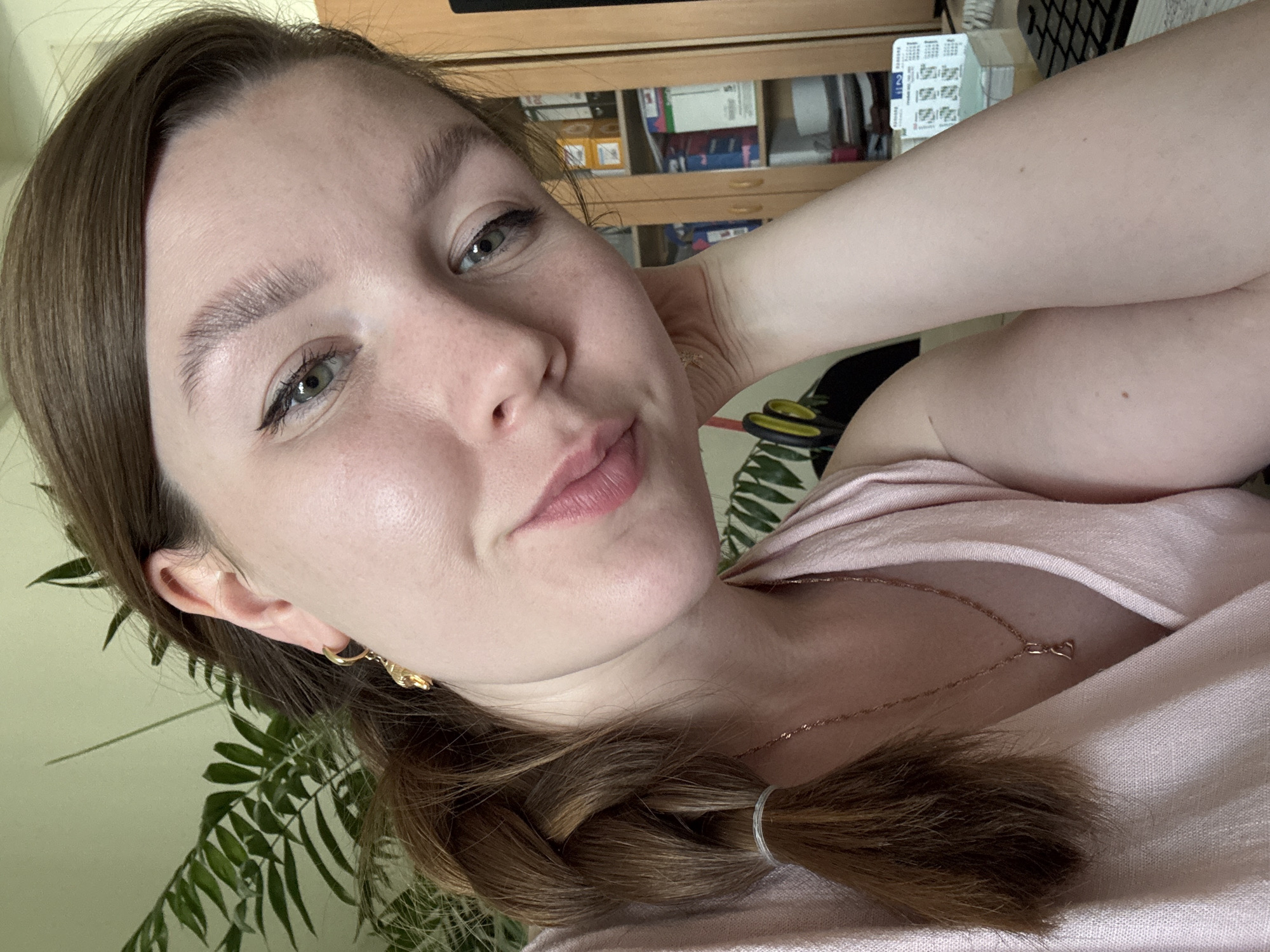 A woman with fair skin and light brown hair braided and secured with a white elastic band is posing for a close-up selfie. She is wearing a light pink top and a delicate gold necklace. Her left hand is raised and resting against her head. She has a neutral expression with a slight smile. She is wearing gold hoop earrings. Behind her is a fern, a wooden cabinet with various items inside, and a glimpse of a computer keyboard. The location is possibly indoors and potentially in Izhevsk, Russia.