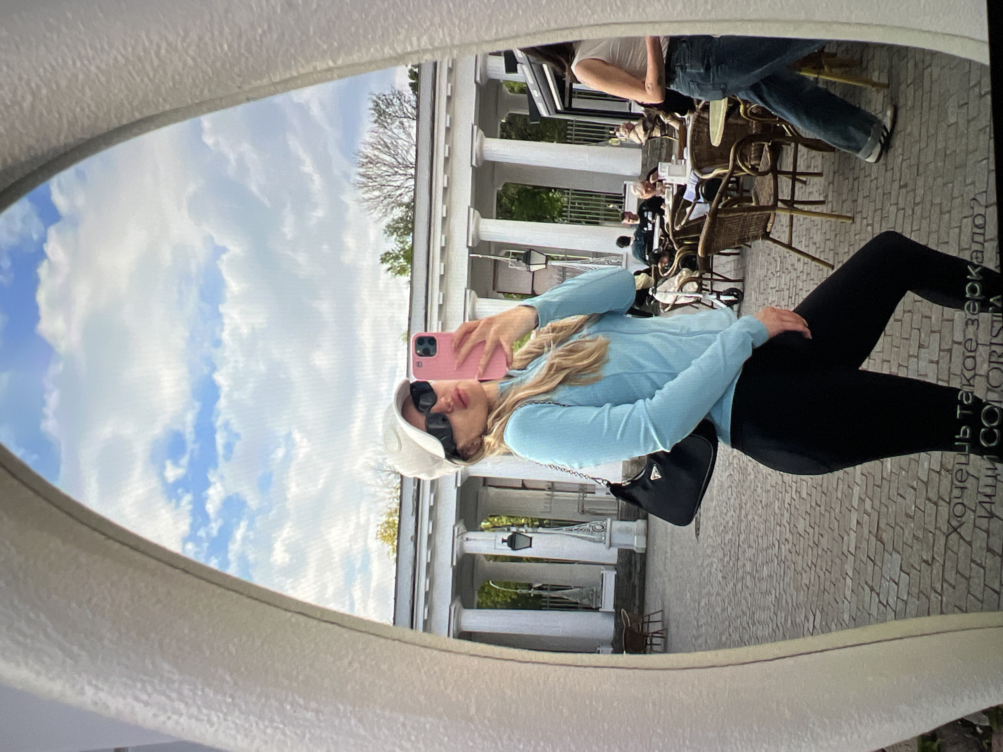 A woman in a blue long-sleeved shirt, black leggings, a white baseball cap, and sunglasses is taking a mirror selfie in front of a neoclassical building with columns. She is holding a pink phone in her right hand. A black Prada handbag is slung over her left shoulder. The background shows an outdoor cafe with wicker tables and chairs, where other people are seated. The ground is paved with cobblestones. The sky is cloudy blue. Text is visible at the bottom of the mirror in Russian, which translates to "Do you want a mirror like this? Find COLORISTA." The location is described as Unknown, Russia.