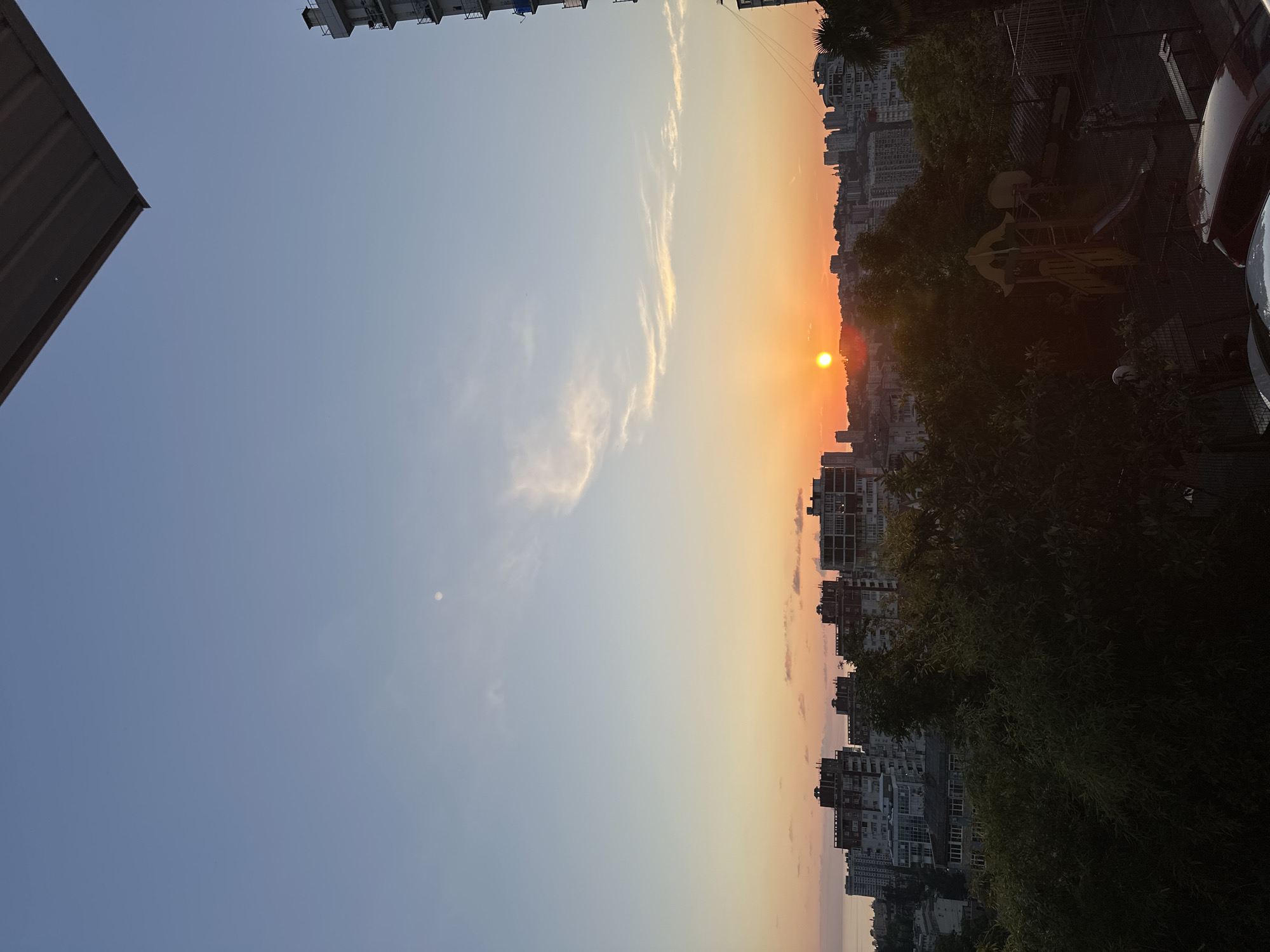 The image captures a sunset over a city that is likely Sochi, Russia, given the distinct architectural styles of the buildings and the presence of palm trees, which are common in the region. The sun, a bright orange orb, is setting near the horizon, casting a warm glow across the sky and illuminating a line of buildings. The sky transitions from a clear blue at the top to soft oranges and pinks near the horizon.
In the foreground, dark green trees obscure part of the view, suggesting a natural setting within or near the urban environment. Beyond the trees, a cluster of multi-story buildings, characteristic of Soviet-era architecture, dominate the landscape. Some of these buildings appear to be residential blocks, while others might be commercial or administrative structures. A playground with colorful equipment is visible among the trees, hinting at a residential area.
The sea is visible on the left side of the image, merging with the horizon. On the right, a tall, modern building under construction or recently completed stands out against the older cityscape. Several cars are parked in the lower right portion of the image, adding a contemporary element to the scene. The overall impression is one of a coastal city at dusk, with a mix of old and new architecture, and a sense of everyday life unfolding as the day ends.