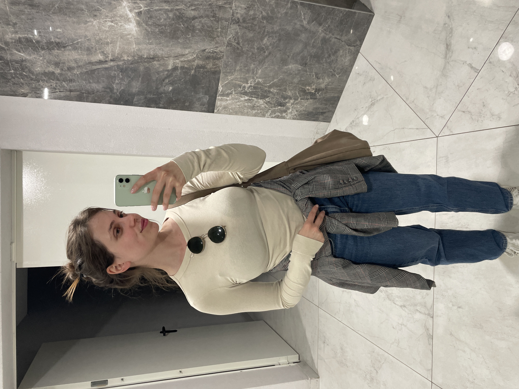 A young woman is captured taking a mirror selfie in an indoor setting. She stands upright, facing the mirror, holding a light green smartphone in her right hand to take the picture. Her hair is styled in a casual bun, and she appears to be looking at the phone screen with a slight, pleasant expression.
She is dressed in a casual yet stylish outfit. She wears a light cream or beige long-sleeved ribbed top, with a pair of dark, round sunglasses hooked onto the neckline. Her lower body is clad in blue denim jeans, and a grey and white plaid jacket is casually tied around her waist. A light brown or beige shoulder bag is visible on her right side, draped over her shoulder.
The background suggests a modern interior space, possibly a hallway or lobby. The floor is composed of large, light-colored, marble-patterned tiles with dark grout lines, forming a geometric design. The wall behind her, above a white baseboard, features large, dark grey marble-patterned tiles that reflect some overhead lighting. To her left (viewer's right), a white door or panel with a dark handle is partially visible. The lighting is bright and even, illuminating the scene clearly. No visible text is present in the image.