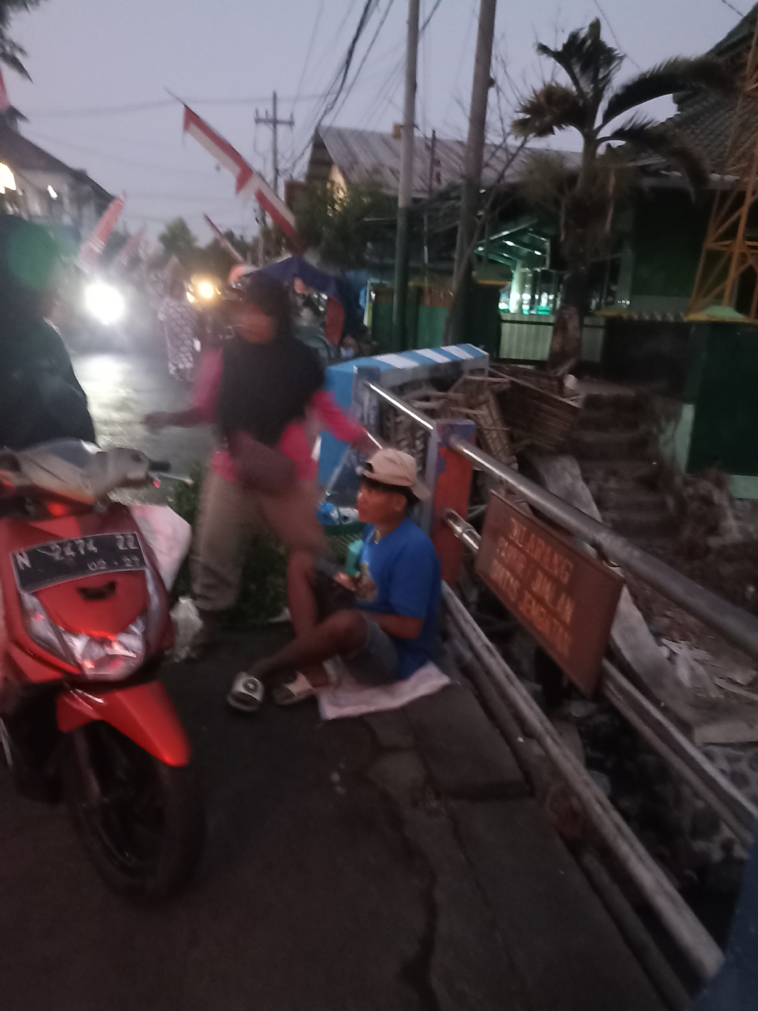 A low-angle, medium shot captures a street scene in Ditotrunan, Indonesia, during what appears to be twilight or early evening. The image is dark and slightly blurred, suggesting a candid, possibly mobile capture.
In the foreground to the left, a red scooter is parked, its front visible with a license plate reading "N 2474 ZZ" and a date "02 . 27". To the right of the scooter, a young boy wearing a light-colored baseball cap and a blue t-shirt sits on the ground, his legs crossed. He is looking towards the right of the frame and appears to be holding something small and dark in his hands.
Behind the boy, a woman in a dark headscarf and a pink top is partially visible, moving from left to right. Another person, also in dark clothing, is positioned behind her. Further back, several Indonesian flags, white and red, are displayed, some attached to poles and others appearing to be flags hanging from buildings. Buildings with roofs are visible in the background, along with trees and utility poles.
A sign with Indonesian text, "DILARANG PARKIR / JALAN BUS TEMPATAN", is attached to a metal railing to the right of the boy. The railing itself appears to be part of a barrier or a drainage canal. Debris and construction materials are scattered along the edge of the railing. The overall environment suggests a residential or commercial street with some ongoing activity.