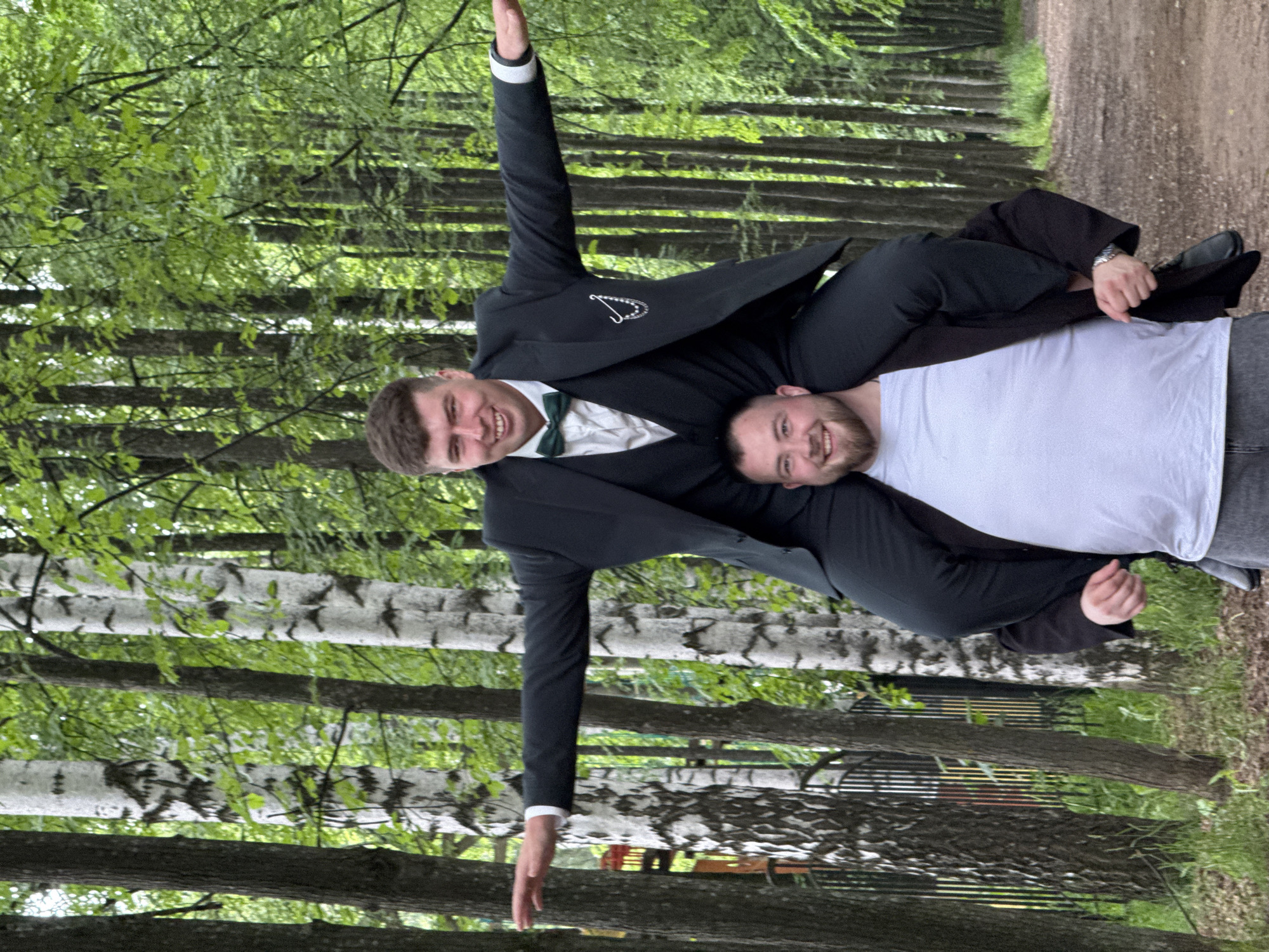 Two men are playfully posing outdoors on a tree-lined path in Chaikovsky, Russia. The image is taken during the daytime, likely on a clear or slightly overcast day, judging by the even lighting and green foliage.
One man, on the left of the frame (when upright), is wearing a dark suit jacket, a white collared shirt, and a dark green bow tie. He has short brown hair and is smiling broadly. He is carrying the second man over his left shoulder in a "fireman's carry" style. This man's left arm is raised high in the air, and a decorative, intricate emblem (possibly initials or a symbol) is visible on the lapel area of his suit jacket.
The second man, being carried, is wearing a white t-shirt and gray sweatpants. He has short brown hair and a beard, and is also smiling, looking towards the camera. His head rests on the first man's shoulder/back, and his left arm is wrapped around the first man's back. A watch is visible on his left wrist.
The background features a clear dirt or gravel path to the right, bordered by numerous trees on both sides. Some trees in the mid-ground have distinctive white, possibly birch, bark with dark markings. The dense green leaves suggest either spring or summer. The overall mood of the image is joyful and amicable, reflecting the men's smiling expressions. No visible text is present in the scene.