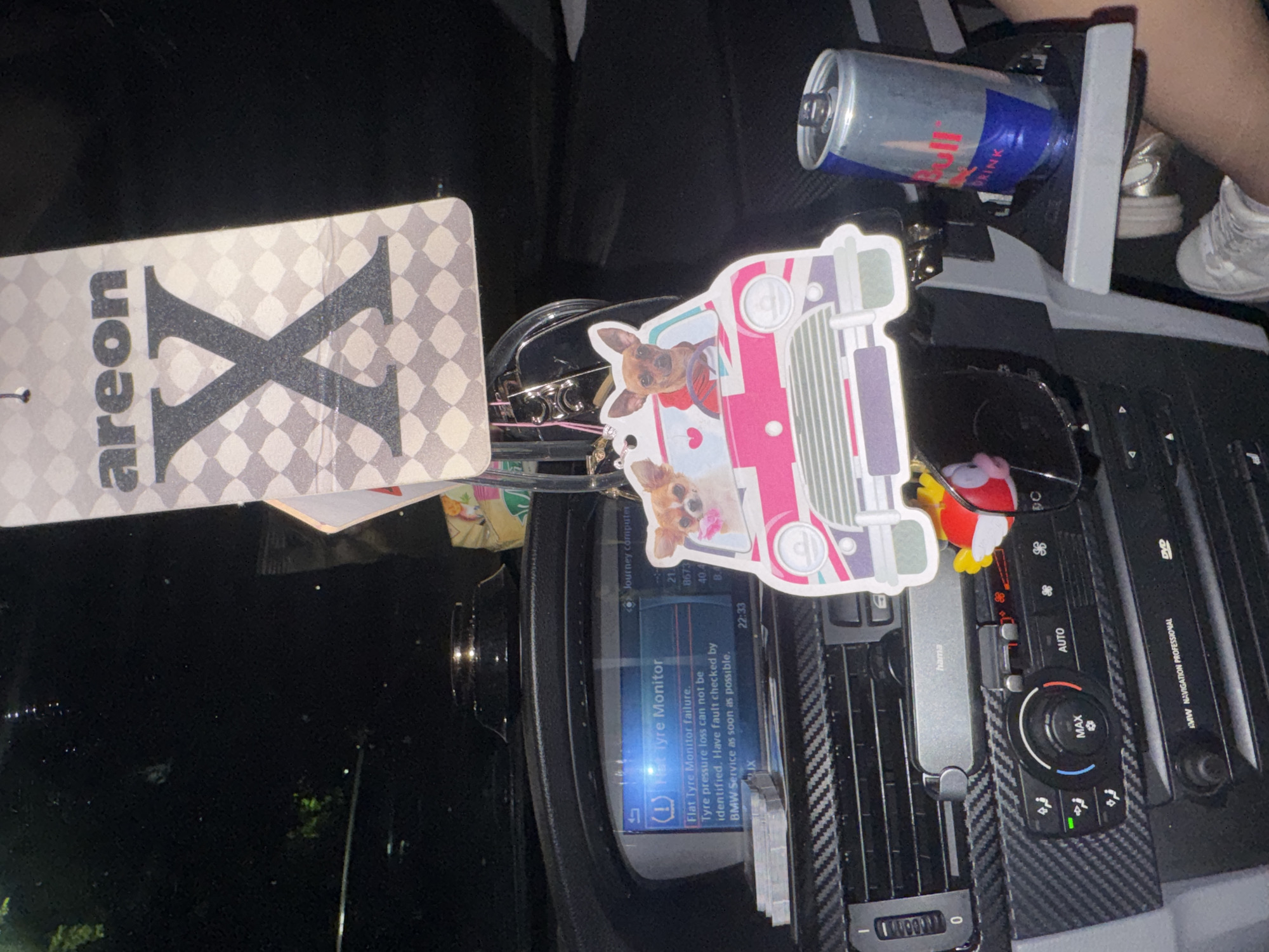 The image captures a detailed view of the dashboard and center console of a car at night, likely in Varna, Bulgaria. The scene is illuminated by the car's interior lights and screens, while the exterior is dark, visible as a blurry black background.
In the upper left, an "areon X" branded car air freshener hangs, featuring a large 'X' on a checkered background. To its right, a collection of items sits on or near the dashboard: a keychain with an illustration of a pink car and two dogs, a small character figurine, and a pair of sunglasses.
A key element of the image is the car's infotainment screen, which displays a "Flat Tyre Monitor failure" message. The text reads: "Flat Tyre Monitor failure. Tyre pressure loss can not be identified. Have fault checked by BMW Service as soon as possible." The time "22:33" is also visible on the screen, along with other data like "21", "8673", "40.4", "B.4", and "Journey computer". This indicates a potential issue with the vehicle's tire pressure monitoring system.
Below the screen, the climate control panel is visible, featuring 'AUTO' and 'MAX' settings and a lit green indicator. The dashboard and air vents are accented with a carbon fiber-like trim. A "hama" branded USB device is plugged into one of the air vents. To the right of the climate controls, a "BMW NAVIGATION PROFESSIONAL" unit with a 'DVD' slot is integrated into the console, confirming the vehicle is a BMW.
In the upper right, a Red Bull energy drink can is secured in a cupholder, and part of a person's legs or feet, wearing light-colored shoes, is visible, suggesting someone is seated in the passenger or driver's seat. The overall impression is a moment captured inside a car during an evening drive, with a notable alert indicating a vehicle fault.