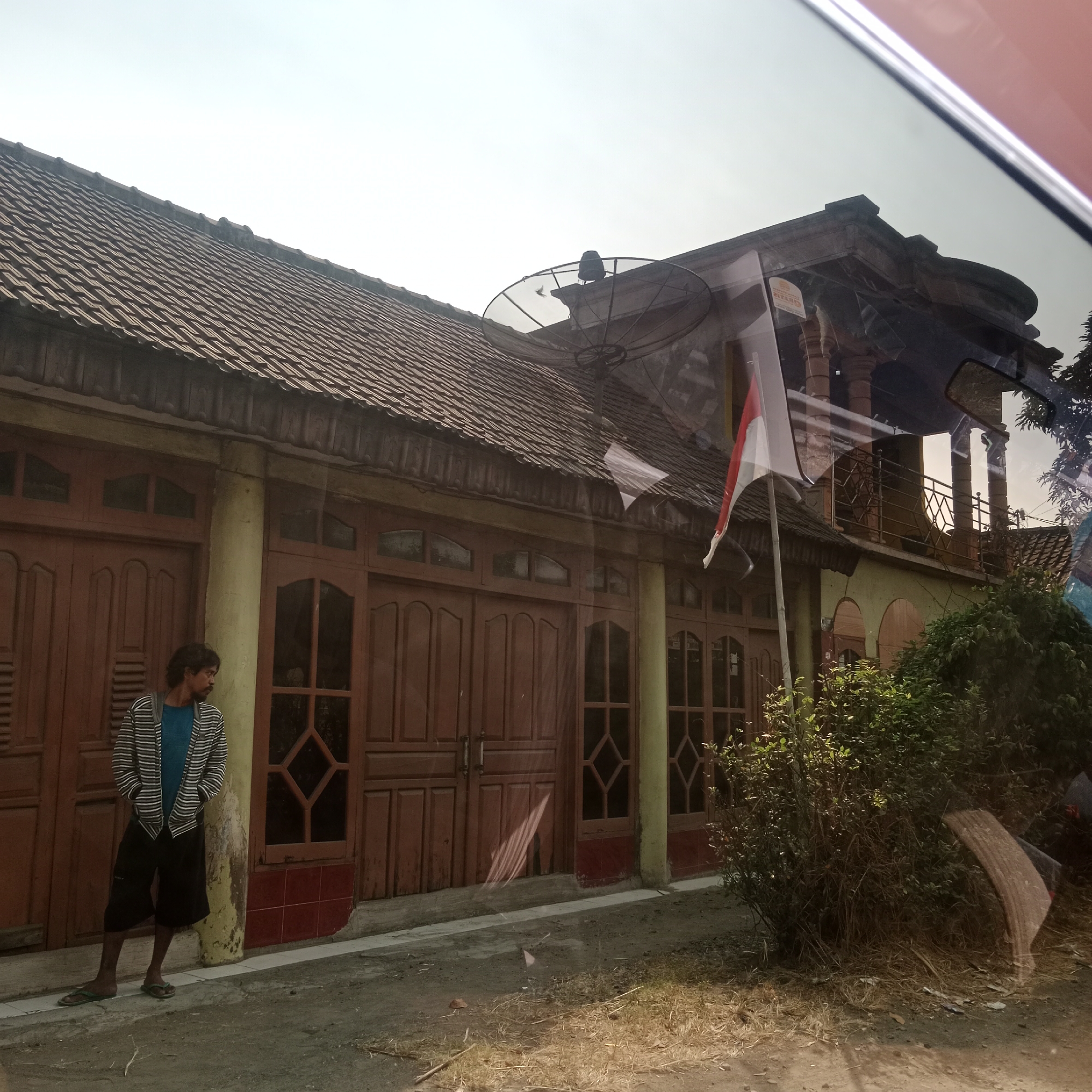 In Gendingan, Indonesia, a man stands to the left of a building with a tiled roof. He wears a striped hooded jacket over a blue t-shirt and black shorts, with green flip-flops on his feet. The building features large wooden doors with diamond-shaped window panes, and a thick, yellowish pillar supports the roofline between two doors. A satellite dish is mounted on the roof. To the right of the building, a lush green bush is prominent, partially obscuring a curved, beige object. An Indonesian flag on a pole is also visible, attached to the building's upper story. The ground in front of the building is a mixture of dirt and dry grass. The scene appears to be captured from a vehicle, as suggested by the reflection of a red car interior and a rearview mirror on the building's facade.