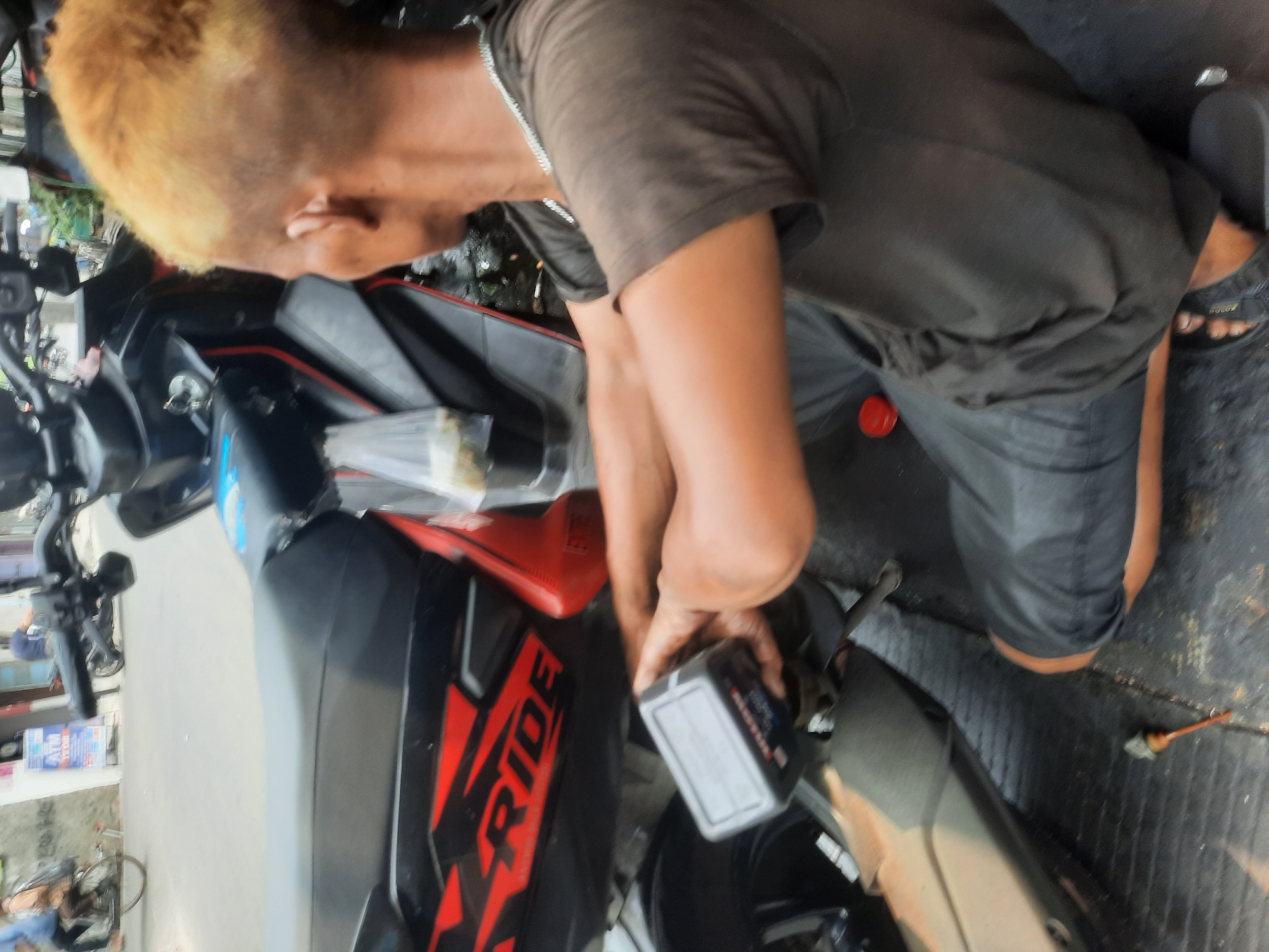 In North Jakarta, Indonesia, a person with bleached blonde hair is working on a motorcycle. They are wearing a dark brown t-shirt and black shorts, with sandals on their feet. The individual is holding a small, gray electronic device in their hands, which appears to be connected to the motorcycle. The motorcycle is red and black, with "RIDE" printed on its side. The scene is outdoors, with a paved surface and some debris on the ground. Other motorcycles and a person on a bicycle are visible in the background, suggesting a street or workshop environment.
