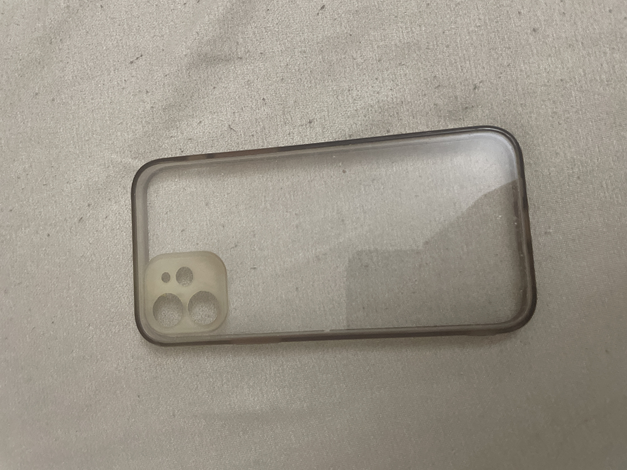 A single, transparent smartphone case is observed lying on a light-colored, woven fabric surface in the city of Ihtiman, Bulgaria. The case is rectangular with uniformly rounded corners. At its upper left, it features a distinct, opaque, off-white raised module designed for a multi-camera system, which contains two larger circular apertures and two smaller circular apertures arranged in a square pattern. The main body of the case is made of clear, flexible plastic, revealing the underlying textile texture and minor surface dust. The edges of the case exhibit a slightly darker, translucent gray tint. The background fabric displays a consistent, finely woven texture with scattered small fibers and minor pilling. No individuals, actions, or additional significant objects are depicted.