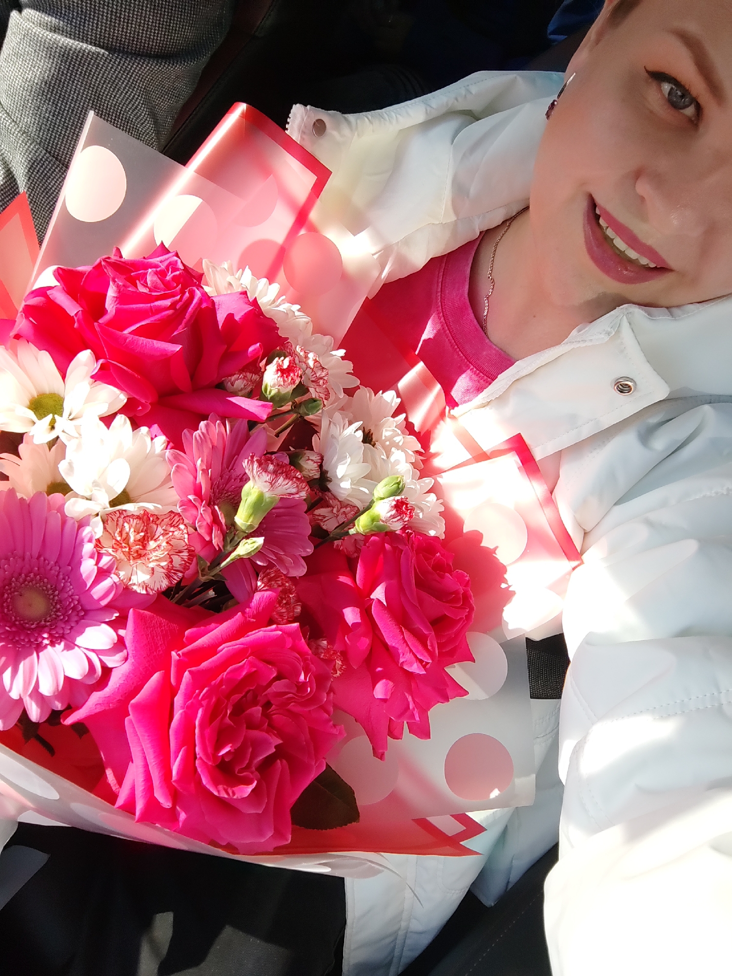 A selfie of a woman smiling with a bouquet of pink and white flowers. The woman is wearing a pink t-shirt and a white jacket. The bouquet contains roses, gerberas, carnations, and daisies. The flowers are wrapped in translucent pink and white polka dot paper. The photo was taken in a car. The location is Chaikovsky, Russia.
