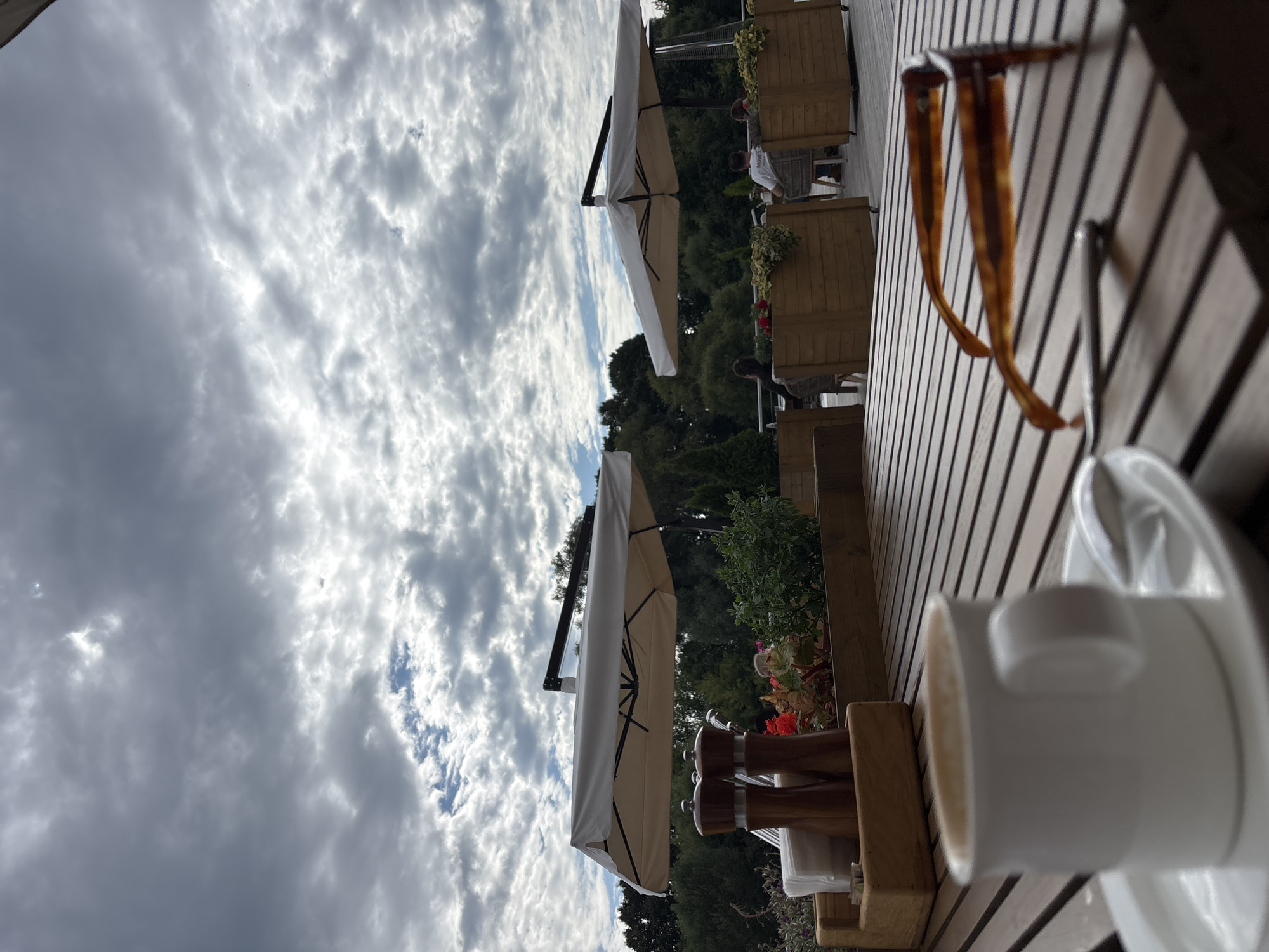 The image depicts an outdoor seating area at a cafe in Zvenigorod, Russia, under a cloudy sky. In the foreground, a wooden table is set with a white cup of coffee on a saucer, a spoon, a pair of tortoise-shell glasses, and a wooden caddy containing salt and pepper grinders and a stack of napkins. Beyond the table, two large white umbrellas provide shade over tables where a few people are seated, some engaged in conversation. Planters with green foliage and red flowers are placed between the tables and the surrounding trees. The overall atmosphere suggests a casual dining experience in a natural setting.
