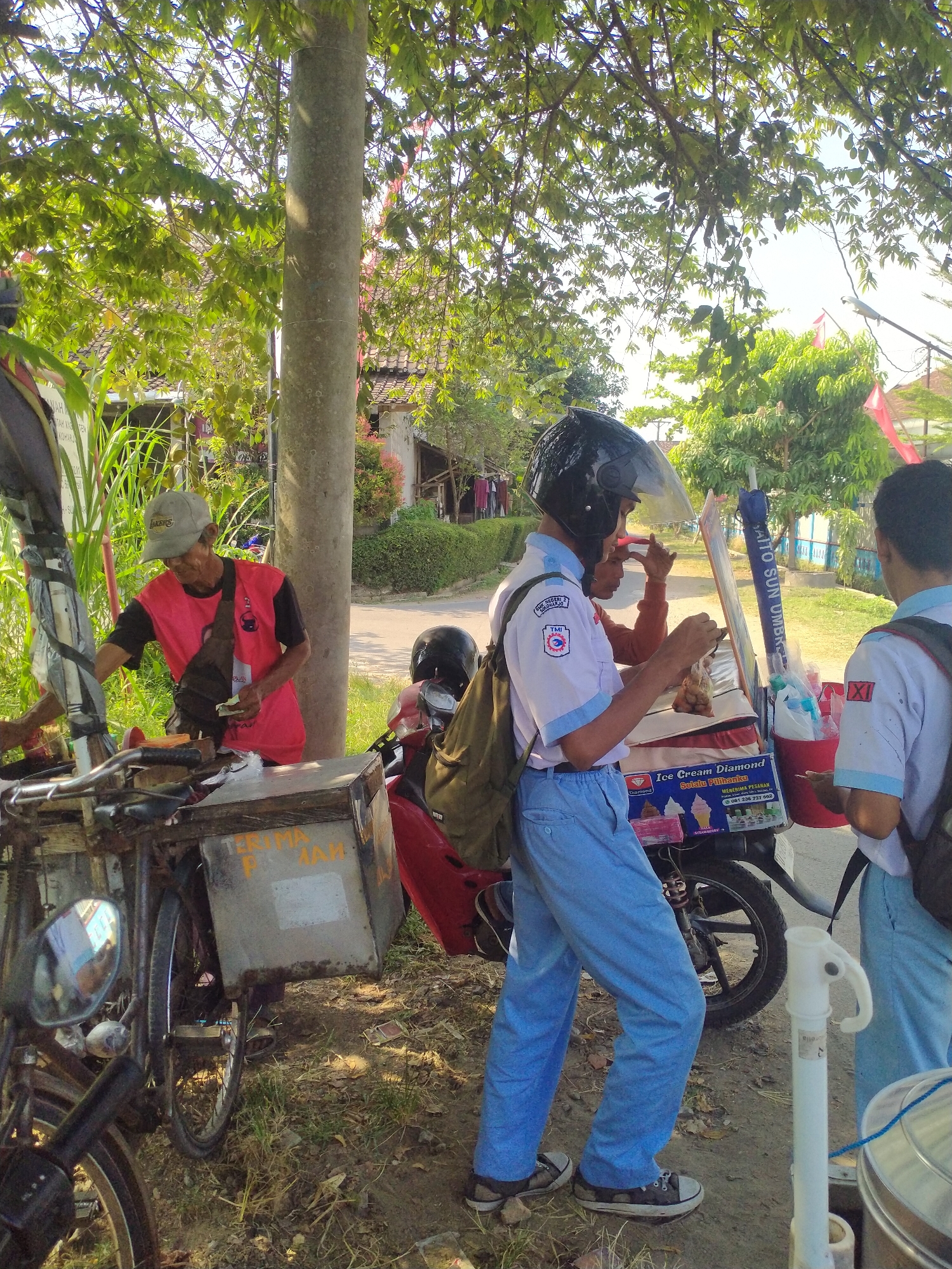 In Begajah, Indonesia, an outdoor scene unfolds near a tree-lined path beside a street. A vendor, wearing a red and black t-shirt and a grey cap, stands by a food cart mounted on a bicycle, which is laden with various items. Beside him, a young person in a white and blue school uniform, complete with a black helmet, is engaged in a transaction at a motorcycle-mounted ice cream cart. Another individual in a similar school uniform stands nearby, observing. The ice cream cart displays an advertisement for "Ice Cream Diamond." The ground is a mix of dirt and sparse grass, with shadows cast by the trees indicating a sunny day. Red flags are visible in the background, suggesting a local event or decoration.