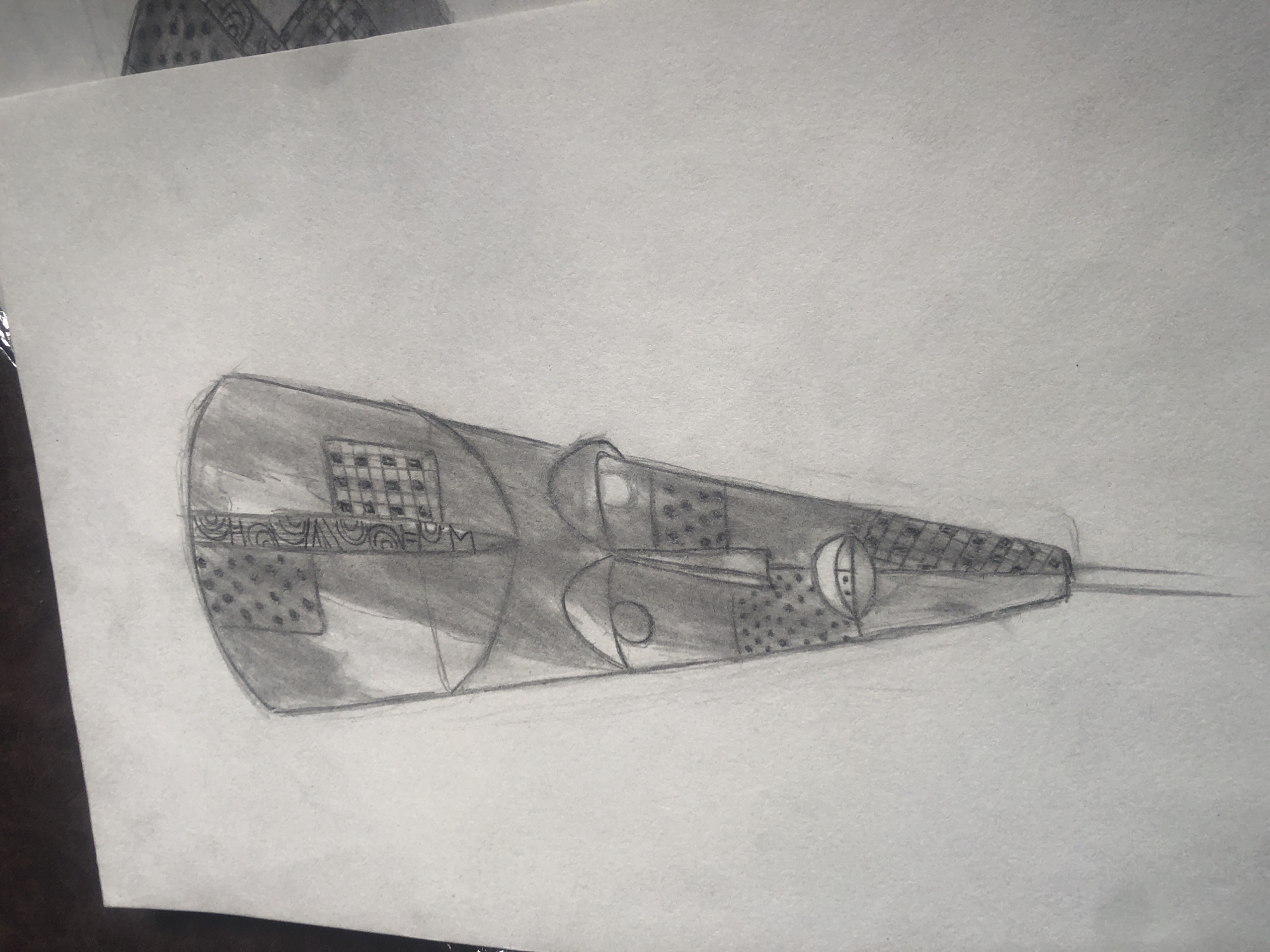 The image is a pencil sketch of a traditional African mask on a textured white paper. The mask is elongated and tapers to a point at the bottom. It has a flattened, rectangular top section with various patterns, including a grid of squares and a section with dots. The face of the mask features two almond-shaped eyes, a triangular nose, and a mouth depicted with simple lines. The surface of the mask is textured with dots and cross-hatching in different areas, suggesting carved designs. The mask is drawn with light and shadow to give it a three-dimensional appearance. The sketch is placed on a surface that appears to be a wooden table, with some other blurred objects visible at the top of the image. The background is a plain greyish-white. The sketch does not contain any elements that would allow for its location in Ado, Nigeria, to be determined.