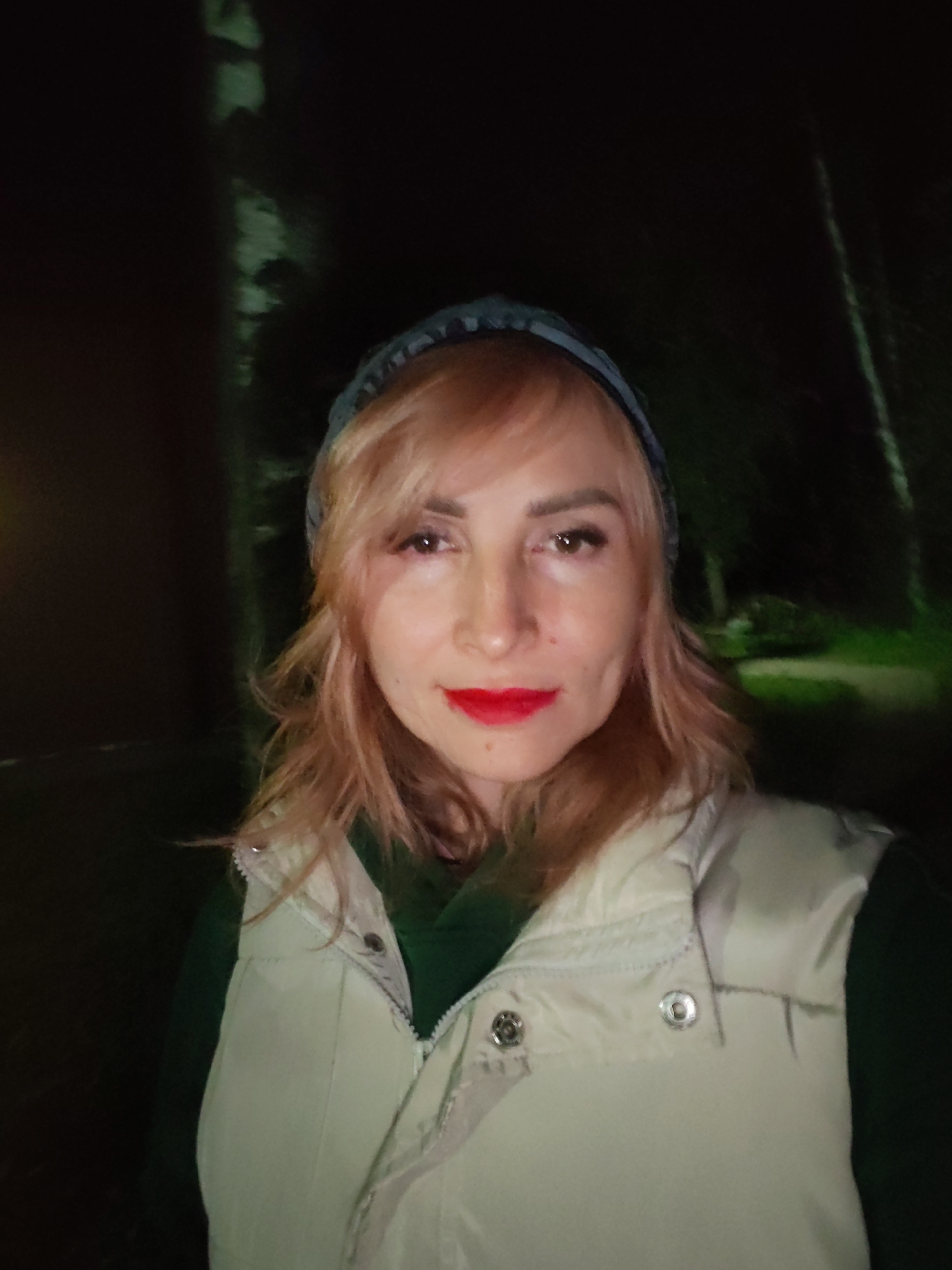 A woman with shoulder-length blonde hair and bright red lipstick poses for a selfie at night, likely in a park or wooded area in Chaikovsky, Russia. She is wearing a light-colored puffer vest over a dark green hooded sweatshirt and a patterned beanie. The background is dark with hints of trees and a faint green glow, suggesting artificial lighting.