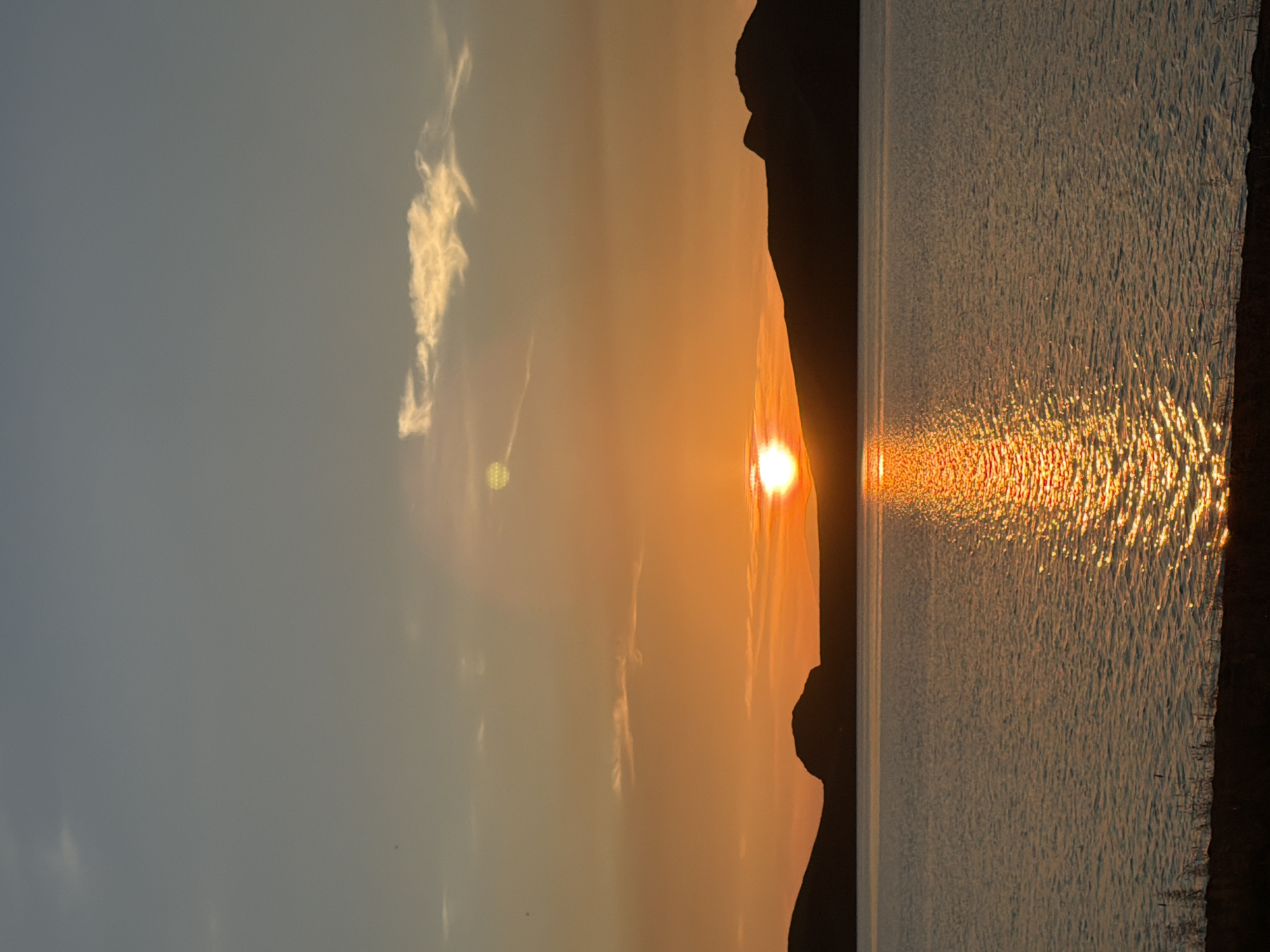 An outdoor vertical shot captures a serene sunset over a body of water, with silhouetted hills in the background. The sky transitions from a pale blue at the top to a vibrant orange near the horizon, a testament to the setting sun. A few wispy clouds are scattered across the sky, one of which appears illuminated by the sun's glow. The sun itself, a bright, white orb, is positioned just above the silhouetted hills, casting a shimmering golden reflection on the water's surface. The water appears calm, with gentle ripples reflecting the warm hues of the sky. The foreground shows a dark, silhouetted shoreline, hinting at a natural landscape. The scene is tranquil and picturesque, evoking a sense of peace and natural beauty. The location is unknown, Russia.