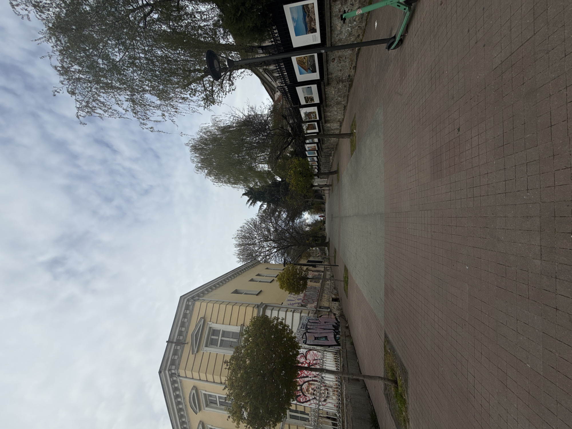 This image captures a street scene in Sofia, Bulgaria, featuring a paved pedestrian pathway, an old building, trees, and an outdoor photo exhibition. The overall impression is of an overcast day.
On the left side of the frame, a multi-story yellow building with classical architectural details dominates, including ornate window frames, cornices, and decorative moldings. The lower section of the building's facade and the metal fence separating it from the path are covered in colorful, though illegible, graffiti. Small, sparsely leafed trees are planted along this side.
To the right, a dark metal fence stretches into the distance, adorned with a series of framed photographs, indicating an outdoor art display. These pictures predominantly feature landscape and nature scenes, each with a white caption underneath, though the text is not decipherable. A black lamppost stands near the fence.
The pedestrian pathway is constructed from square pavers, primarily brown with some sections of grey, leading further into the background. A green electric scooter, displaying the number "5" on its front, is parked against the fence on the right.
The sky is a pale grey, filled with clouds, suggesting an overcast day with muted light. Many of the trees in the scene are bare or have minimal foliage, typical of late autumn, winter, or early spring. No people are visible in the scene.