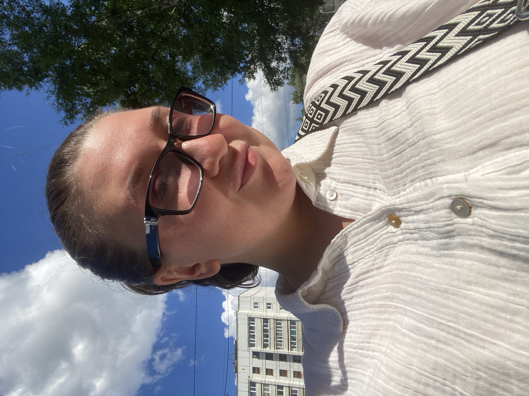 In a close-up, waist-up selfie taken outdoors, a young Caucasian woman with fair skin and dark hair pulled back is looking towards the right side of the frame. She is wearing black-rimmed glasses and a loose-fitting, textured white shirt with buttons. A black and white patterned strap, likely from a bag, is visible across her chest. The background reveals a bright blue sky with scattered white clouds, a tall, multi-story apartment building with numerous windows, and the green foliage of a tree. The lighting suggests a sunny day. This scene is consistent with an outdoor urban environment in Chaikovsky, Russia, likely a residential area with apartment buildings and surrounding trees.