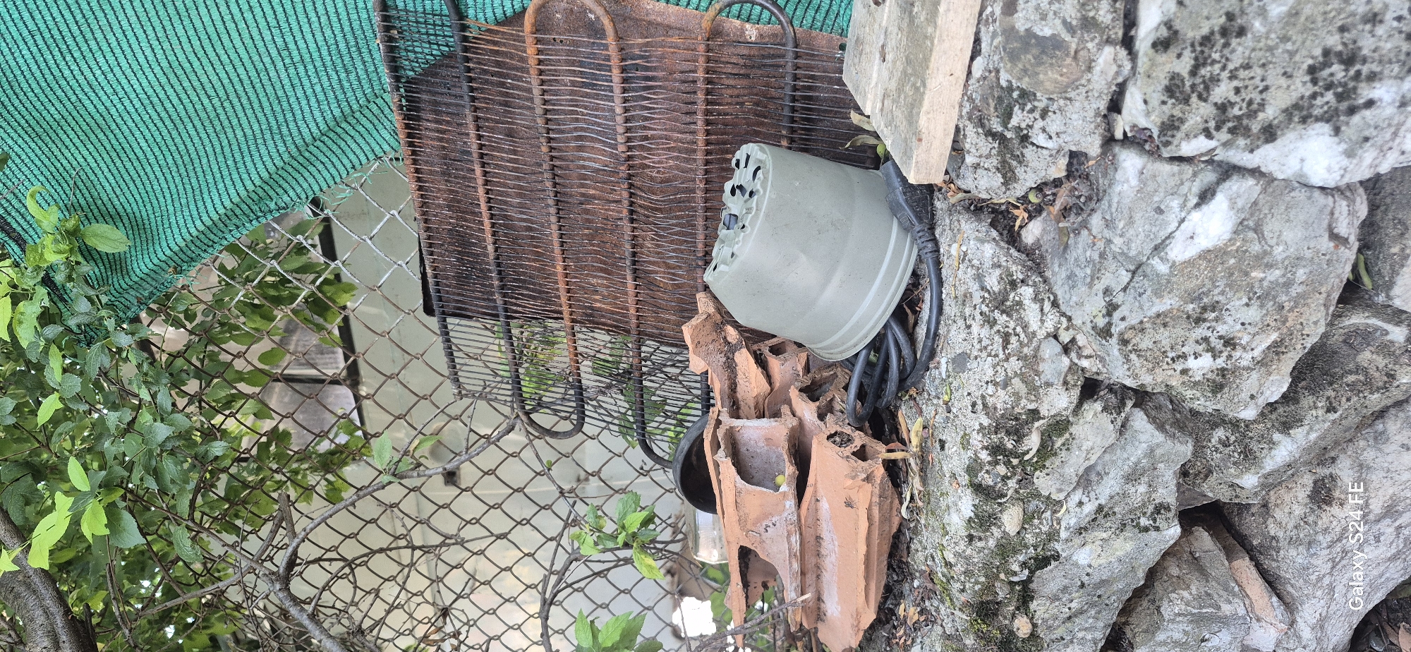 The image depicts an outdoor scene in Malko Tarnovo, Bulgaria, featuring a collection of discarded items piled against a stone wall and chain-link fence. In the foreground, rough-hewn stones, some covered in moss, form a base for the debris. Prominently displayed is a broken terracotta object, possibly part of a roof tile, and a green plastic plant pot lying on its side. A black electrical cord is also visible, coiled near the pot. Behind these items, a rusty, grid-like metal structure, possibly from an old appliance like a refrigerator, leans against the fence. A green mesh fabric is draped over the fence and metal structure. The background shows a chain-link fence, overgrown with green foliage from a tree, with a building or structure visible beyond. The overall scene suggests an area of neglect or an outdoor storage space for unwanted items.