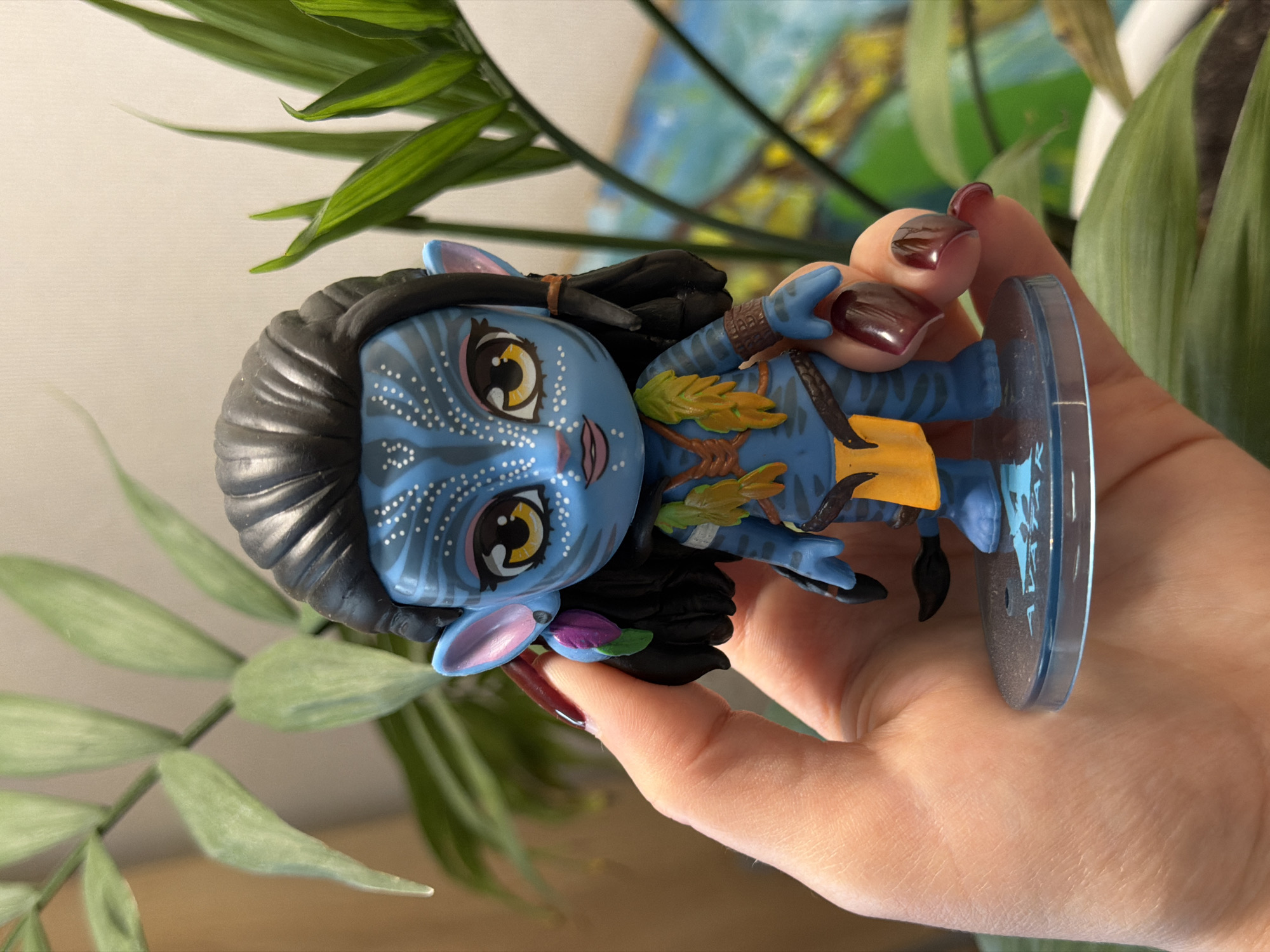 A person holds an Avatar Funko Pop figure. The figure is blue with black hair, yellow eyes, and wearing a yellow loincloth. The person's hand is visible with maroon nail polish. A green plant is in the background. The location is Засечное, Russia.