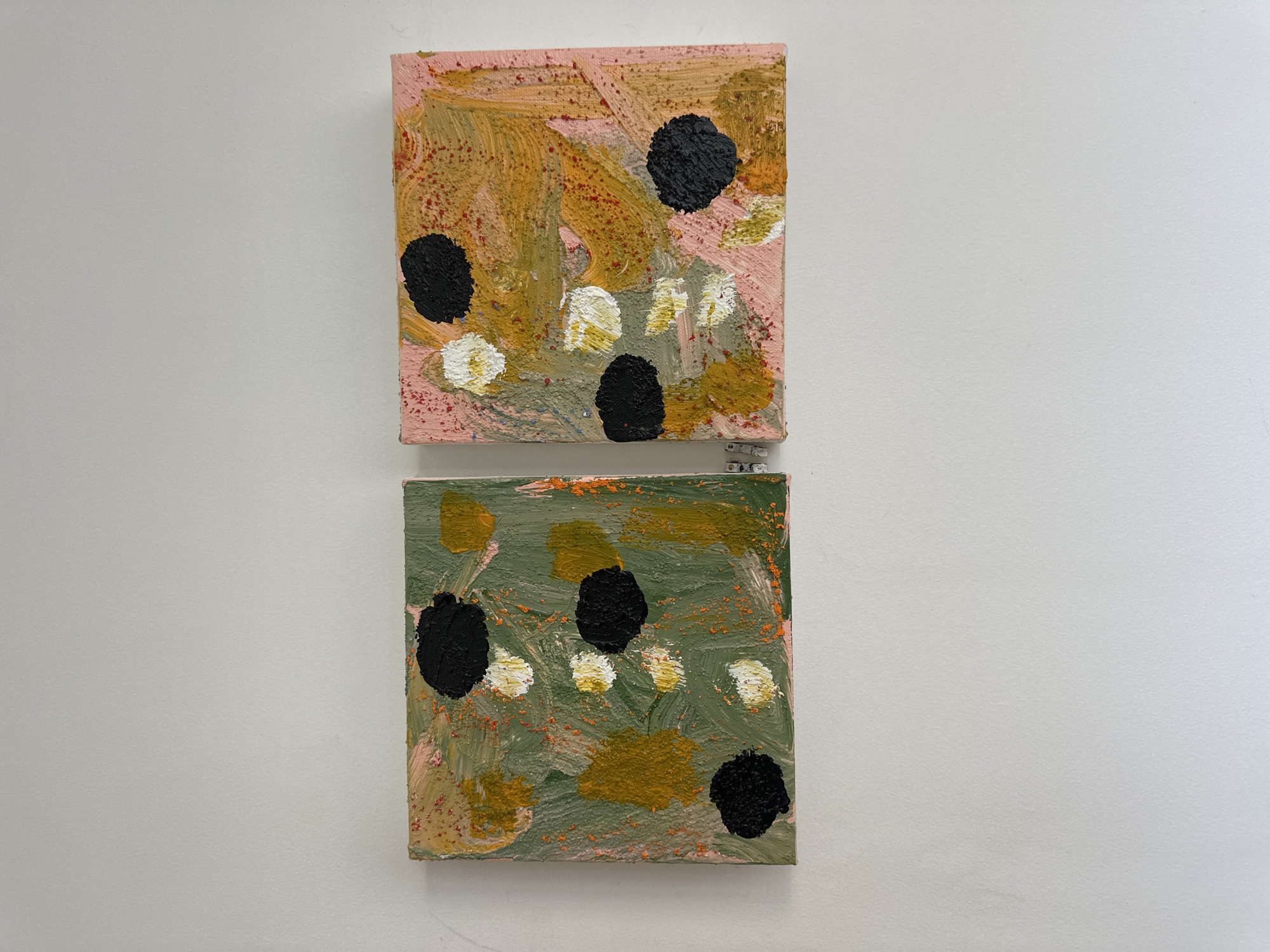 Two small, square abstract paintings hang vertically on a white wall. The top painting has a pinkish-beige background with strokes of gold, small red specks, and four black circular shapes. Patches of white and gold are also scattered across the surface. The bottom painting features a green background with similar gold strokes, red specks, white and gold patches, and black circular shapes. The paintings are likely located in a gallery or building within the Folkestone and Hythe District, United Kingdom.