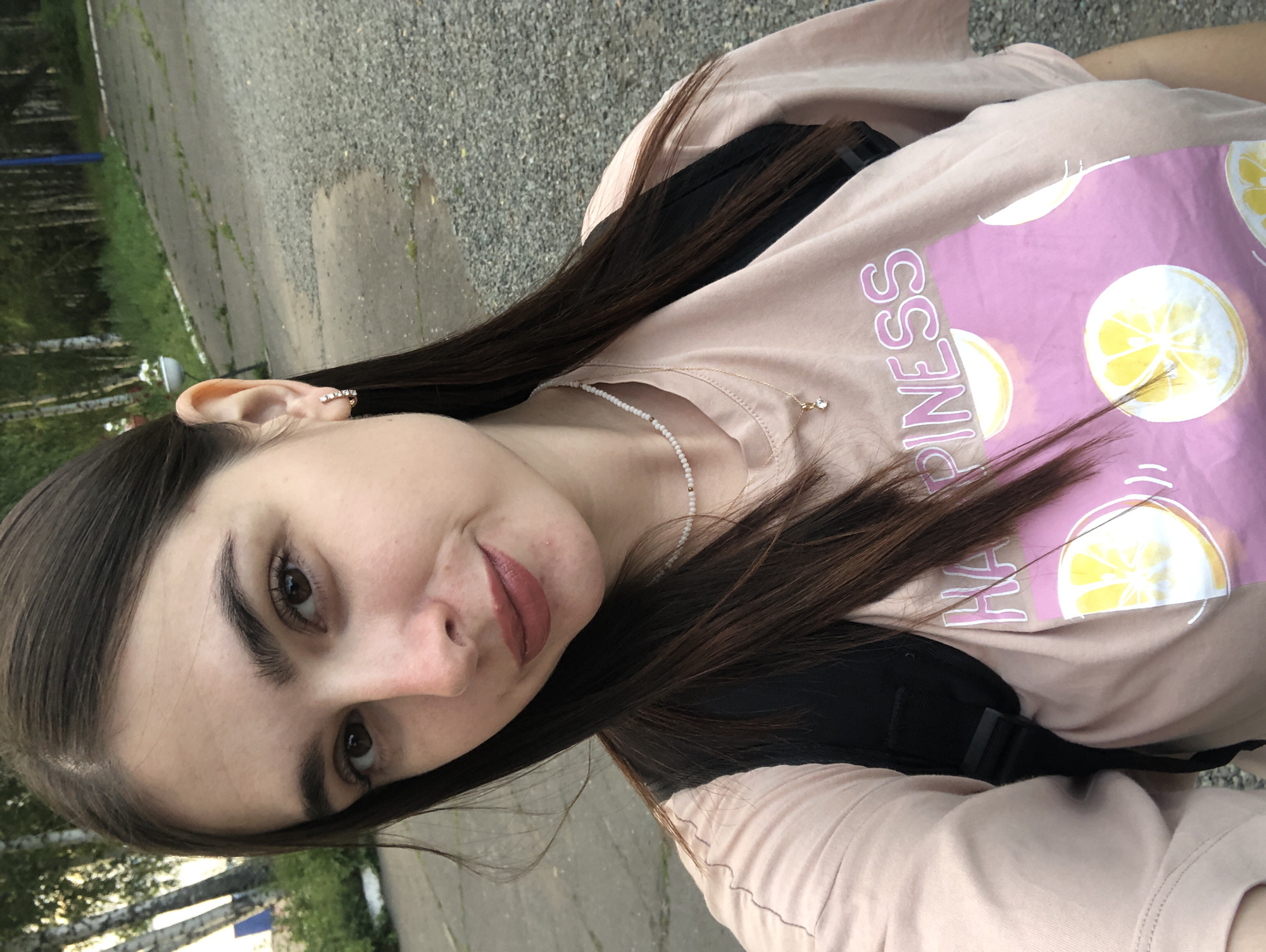 A young woman with long dark hair is pictured in a selfie outdoors. She is wearing a light pink t-shirt with a graphic design of lemons and the word "HAPPINESS" printed on it, and a backpack. She has on a necklace and small hoop earrings. The background suggests an urban or park setting with paved walkways and trees. The image appears to have been taken during daylight hours. The scene is likely located in Chaikovsky, Russia, given the context of the prompt.