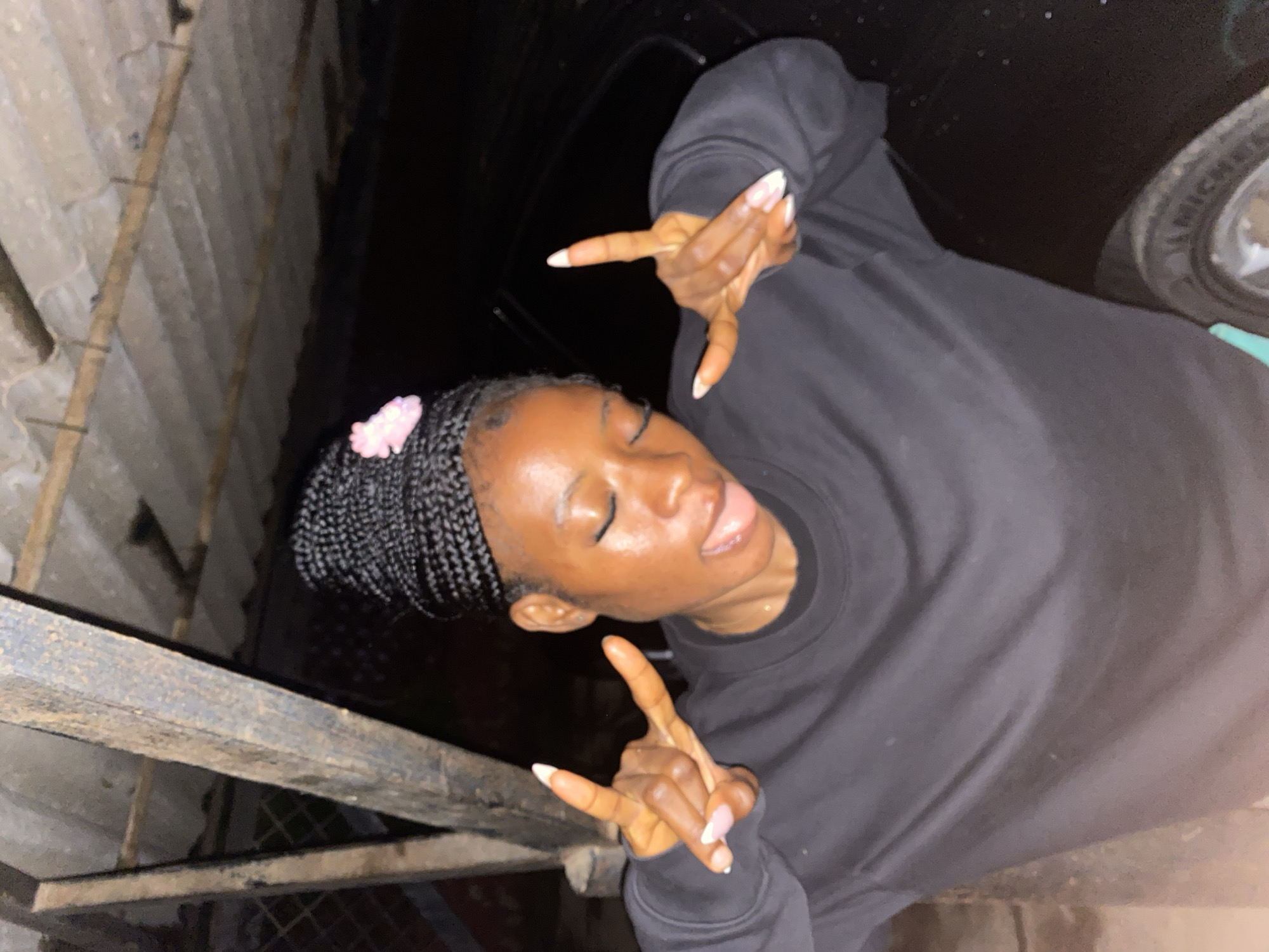 A young Black woman with braided hair and a pink bow is sticking her tongue out and making peace signs with both hands. She is wearing a black sweatshirt. The background is dark, but appears to be an outdoor, possibly urban, setting with corrugated metal roofing and wooden beams visible overhead, and a car partially visible to her right. The overall scene is likely within the city of Jos, Nigeria, given the context provided, but specific landmarks are not discernible.