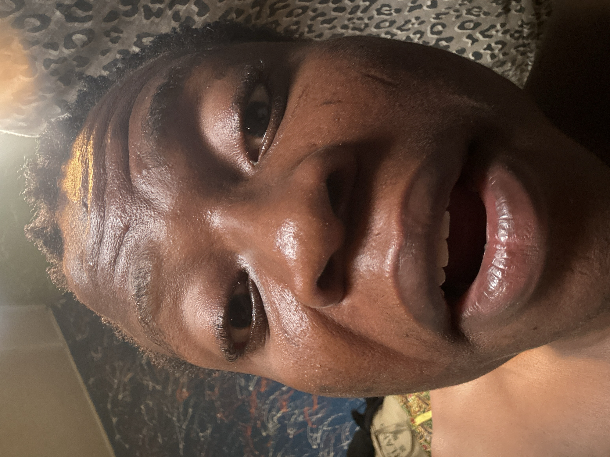 A close-up portrait shows a person with dark skin, likely male, positioned diagonally as if lying down or reclining. The individual is smiling broadly, with their mouth open, revealing white teeth and a pinkish-red tongue. Their dark eyes are open and looking towards the viewer, conveying an expression of happiness or amusement. Short, dark, curly hair frames the face. The skin has a noticeable sheen or oiliness, with visible pores and some minor texture.
A bright light source illuminates the left side of the face, casting prominent highlights on the forehead, nose, and lips. The background features patterned fabrics: a leopard print or similar design behind the head, and a darker, possibly blue/black, patterned material below the face. The person's bare shoulder is visible in the lower right portion of the image. A light-colored object, possibly footwear or part of a blanket, with blurry text that may include "LA" or "TLA," is partially visible in the bottom right corner. The setting appears to be an indoor environment, such as a bedroom or living space.