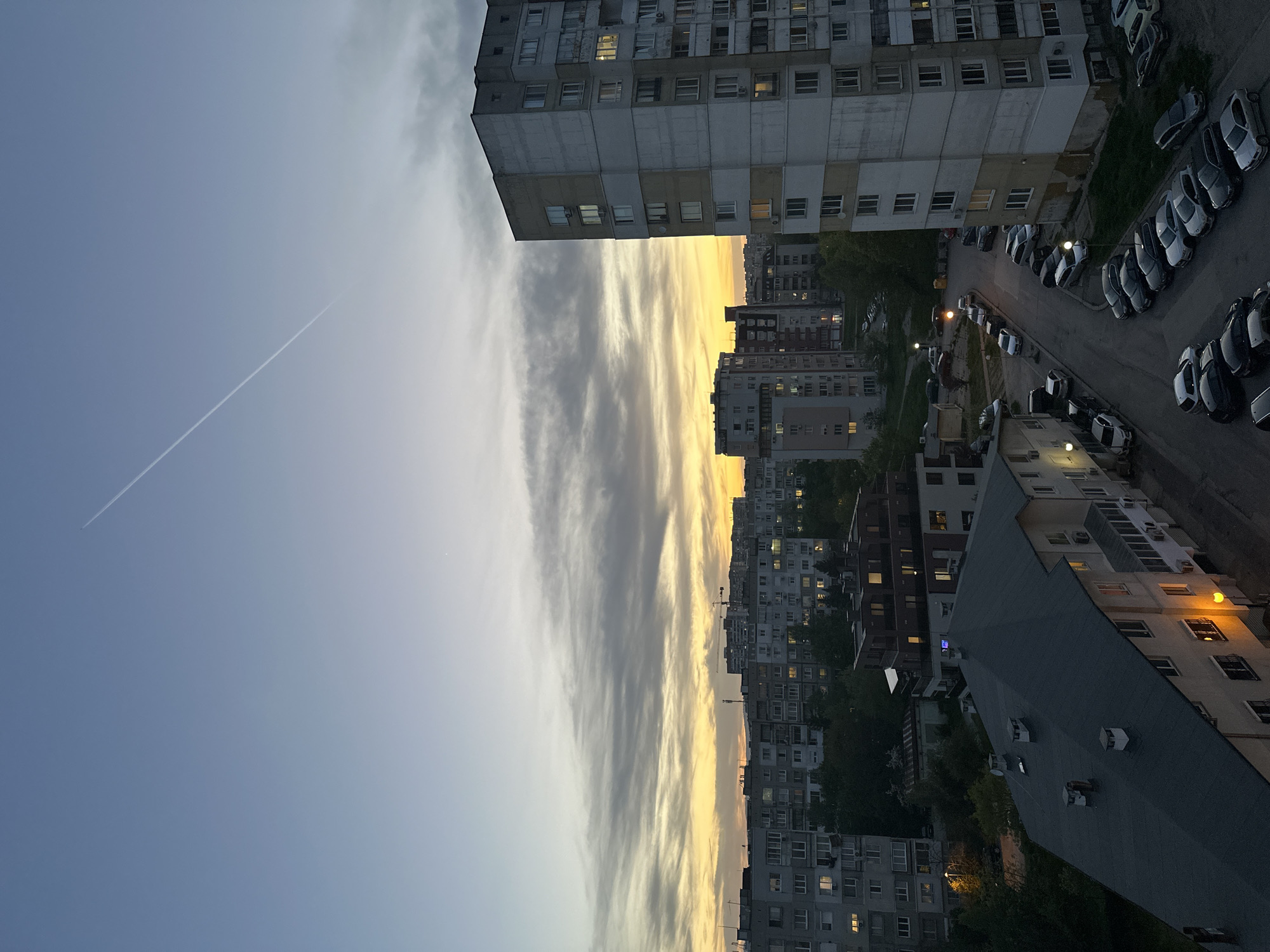 This image, originally oriented vertically but depicting a horizontal scene, presents an urban landscape at dusk or early evening in Sofia, Bulgaria.
The upper portion of the scene is dominated by the sky, which transitions from a clear, light blue on the left to warm orange and yellow hues near the horizon on the right, indicative of a sunset. Streaky, elongated clouds stretch across the middle of the sky, catching the colors of the setting sun. A faint white contrail from an airplane is visible in the upper left section of the sky.
Below the sky, numerous multi-story apartment buildings define the urban environment. The architecture suggests a style common in Eastern European cities, with concrete facades and many rectangular windows. A prominent, tall residential building stands on the right side of the foreground, its windows, some illuminated, hinting at ongoing domestic life. Further into the distance, a dense array of similar buildings extends towards the horizon.
At street level, below the buildings, paved roads and parking areas are visible. Numerous cars are parked along the streets and in designated spots. Several streetlights and lights emanating from building windows are illuminated, confirming the early evening time. Small patches of trees and vegetation are interspersed among the concrete structures.
No people or explicit activity are directly visible, but the lit windows and parked cars suggest the rhythm of city life. The overall mood is quiet and atmospheric, capturing the daily transition from day to night. There is no visible text in the image.