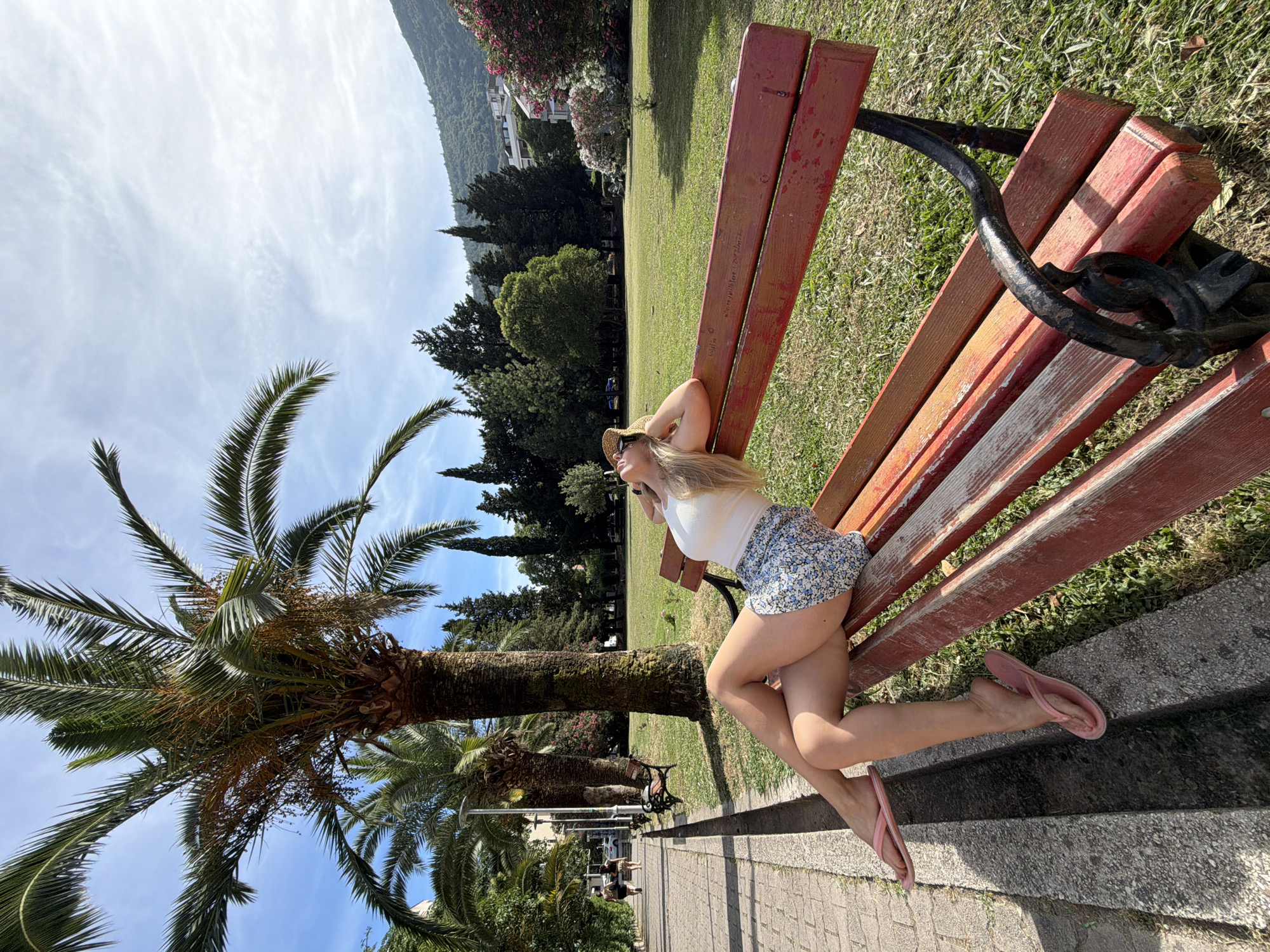 In Budva, Montenegro, a woman wearing a white top and a floral skirt sits on a red park bench in a grassy area. She is leaning back with her head resting on her arm, looking upwards. Her legs are crossed, and she is wearing pink flip-flops. A palm tree is visible behind her, and a paved walkway runs along the left side of the image, with other people walking in the distance. The background shows a lush green lawn, more trees, and a hillside with buildings and vegetation. The sky is blue with scattered clouds.