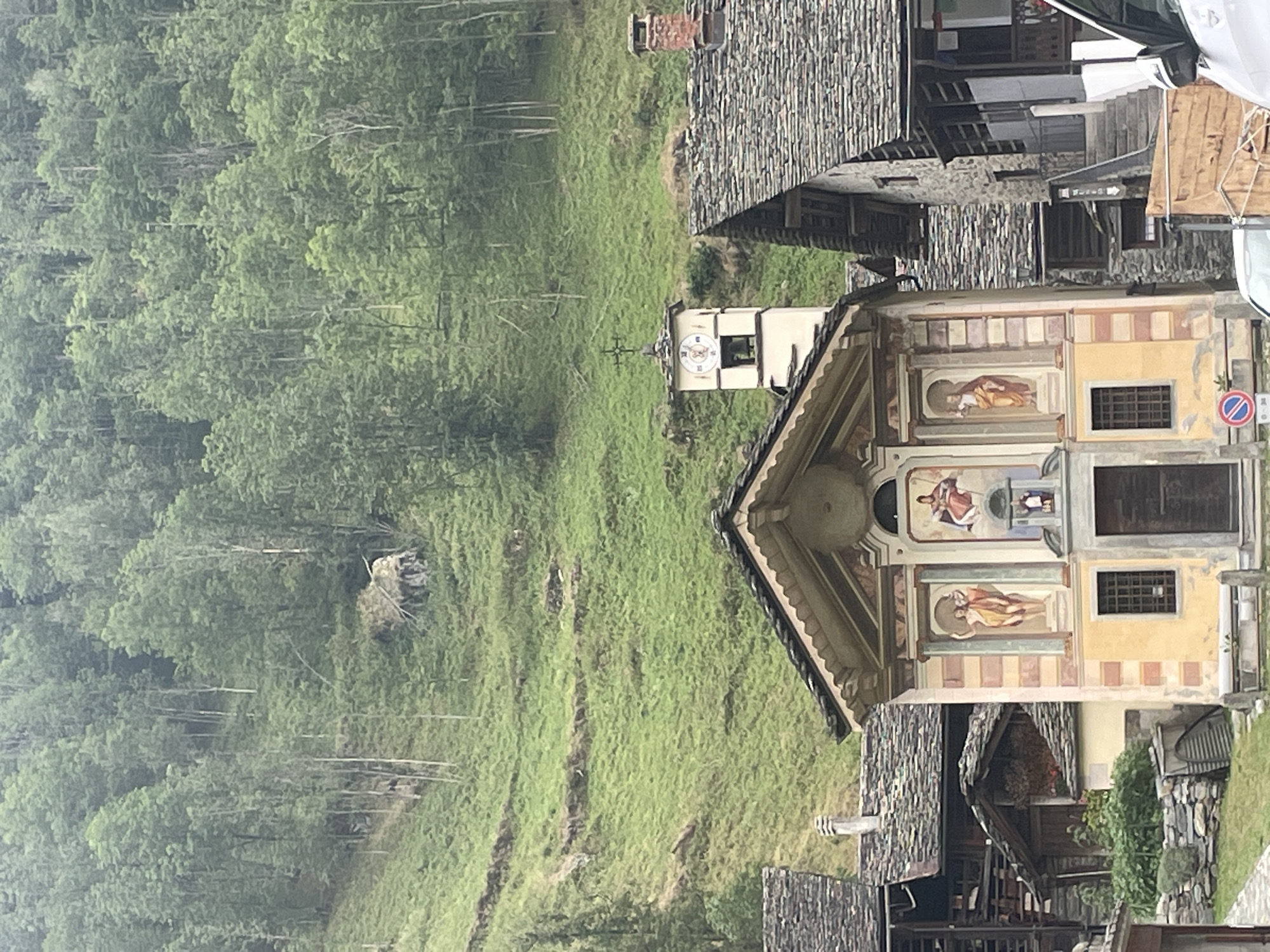 The image shows a quaint village nestled in the mountains of Alagna Valsesia, Italy. In the foreground, a small church with a yellow facade stands prominently. The church features religious frescoes on its exterior, and a bell tower with a clock rises beside it. The architecture of the surrounding buildings, with their slate roofs and stone walls, is typical of the Walser style. Behind the village, a lush green mountainside is covered with dense forest. The terrain is steep, with terraced fields and some evidence of past landslides or debris piles. The overall scene evokes a sense of peaceful seclusion and natural beauty.