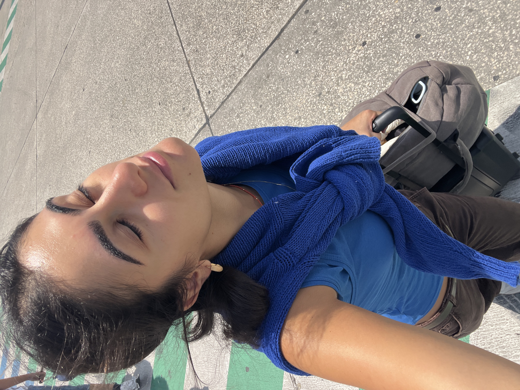 A young woman with dark hair and closed eyes is taking a selfie outdoors. She is wearing a blue tank top, a blue sweater draped over her shoulders, and brown pants. She has a gold earring in her left ear. She is holding the handle of a dark gray duffel bag with her right hand, which is resting on top of a black suitcase with wheels. The ground is paved with gray concrete and has some green markings. The lighting suggests it is daytime. No specific landmarks of Moreira, Portugal are visible.